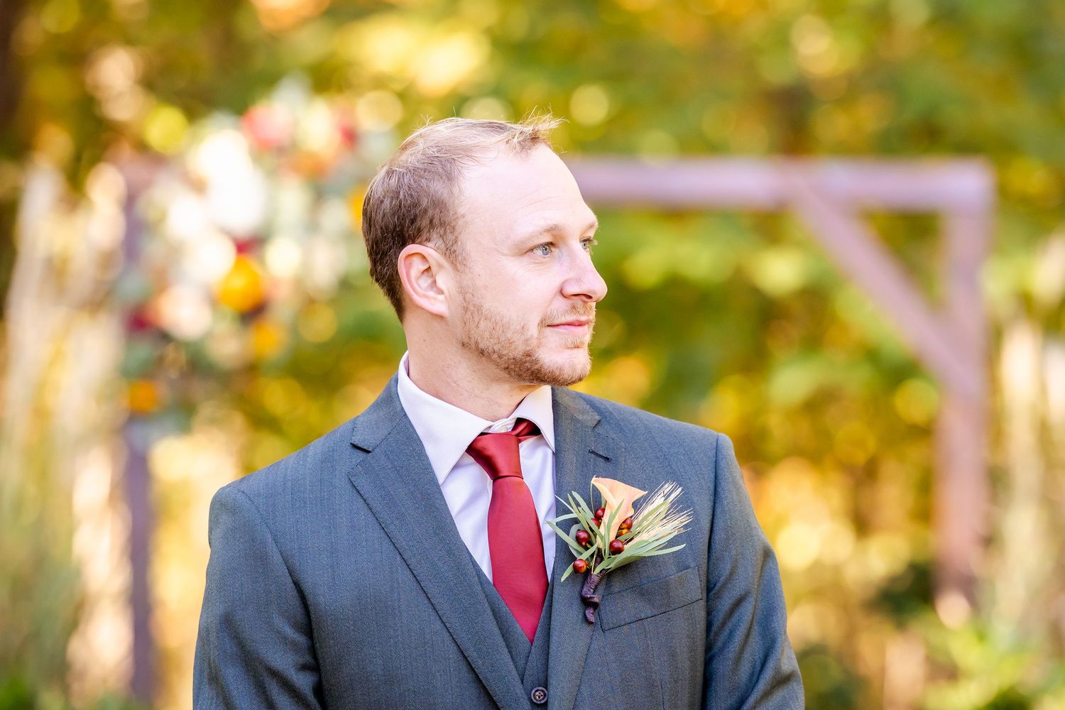 A groom in a navy suit with red tie smiles during an outdoor autumn wedding ceremony.