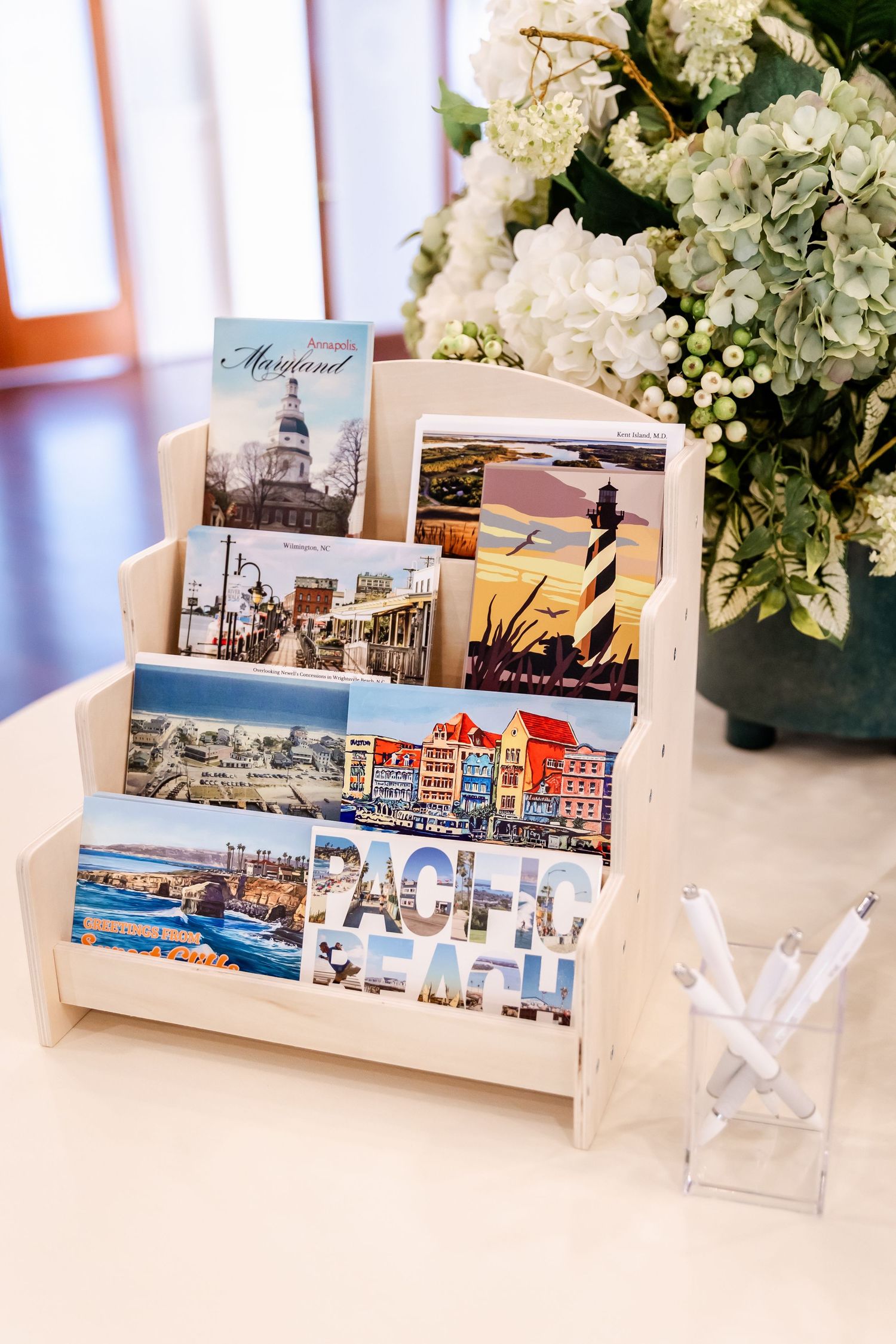 Wedding display holder with postcards of Maryland landmarks and lighthouse images next to white hydrangeas.