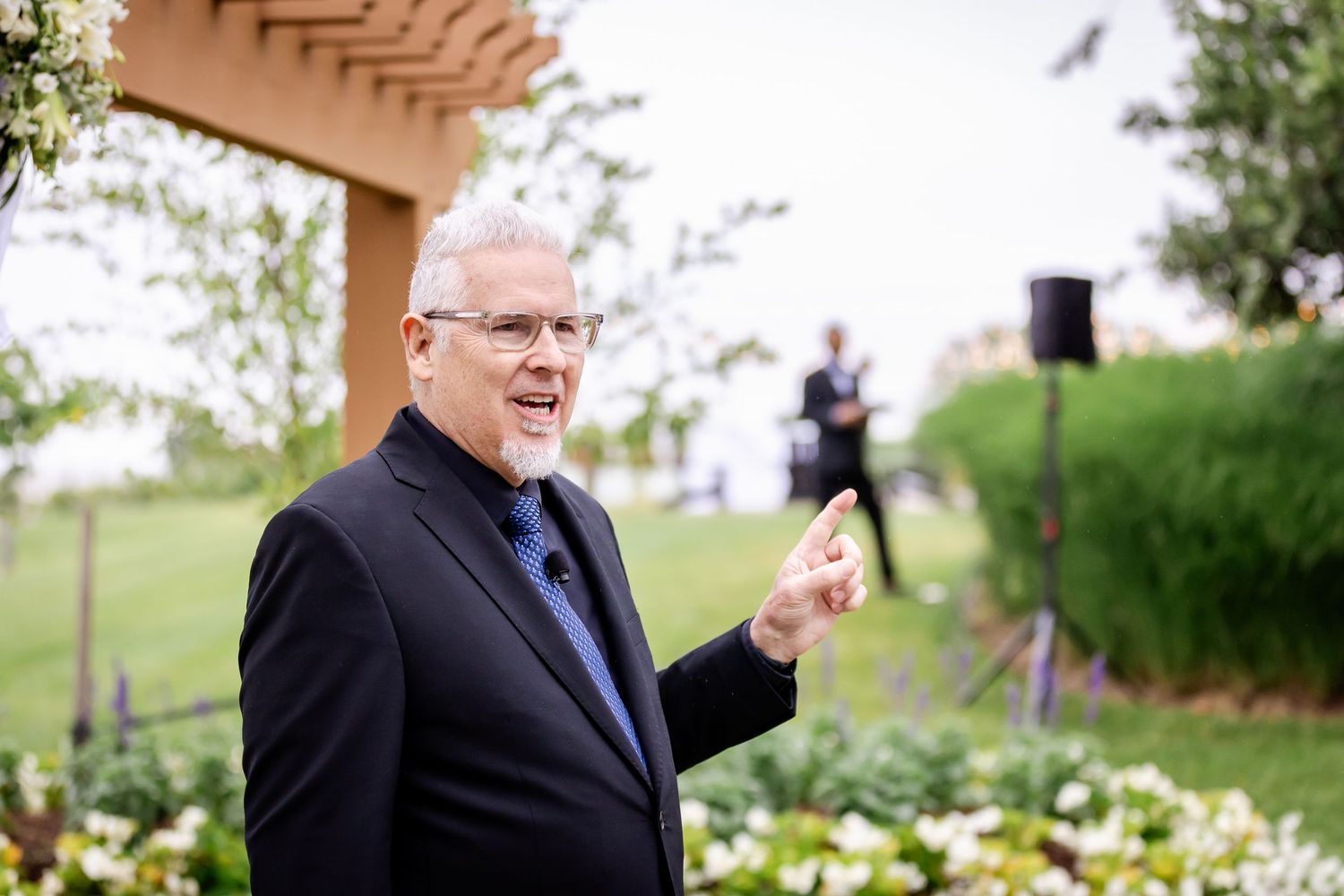 A speaker in a dark suit gestures while giving a speech outdoors in a garden setting.
