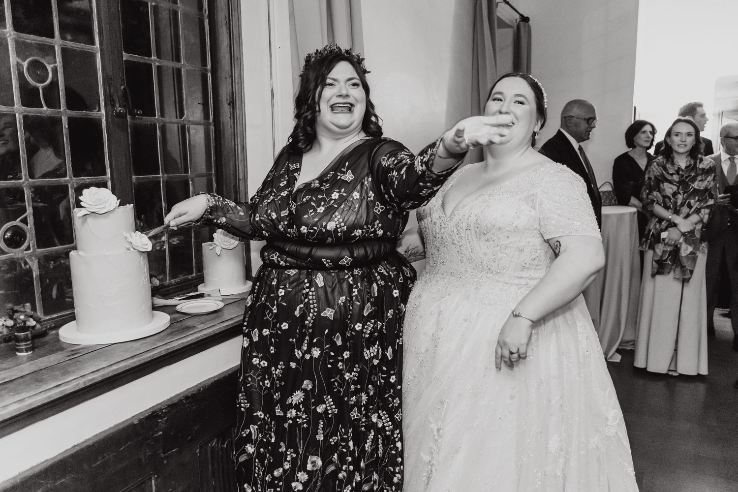 Two women in formal dresses share a laugh at a wedding reception by the cake table.