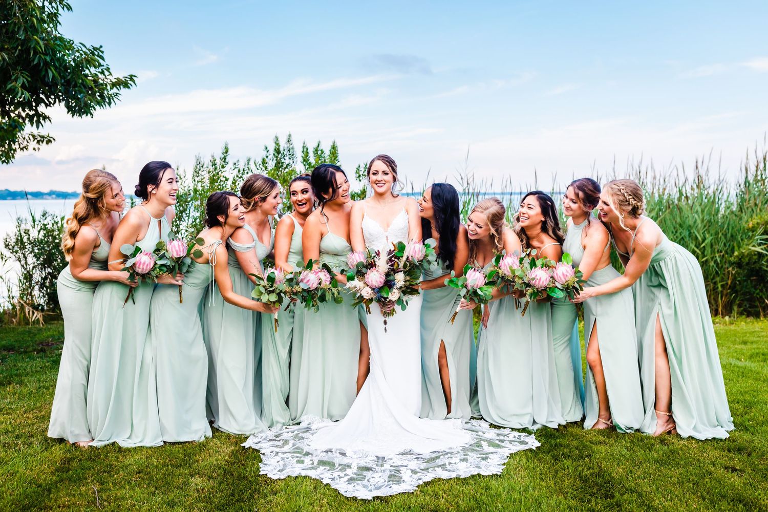 A bride and bridesmaids wearing seafoam green dresses pose together outdoors by a lake on a sunny summer day.