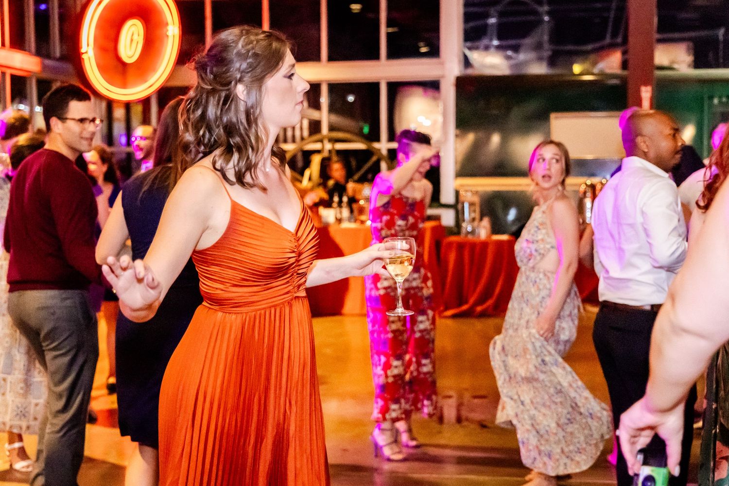 Wedding guests dancing at a reception with one wearing a vibrant orange pleated dress on a lively dance floor.
