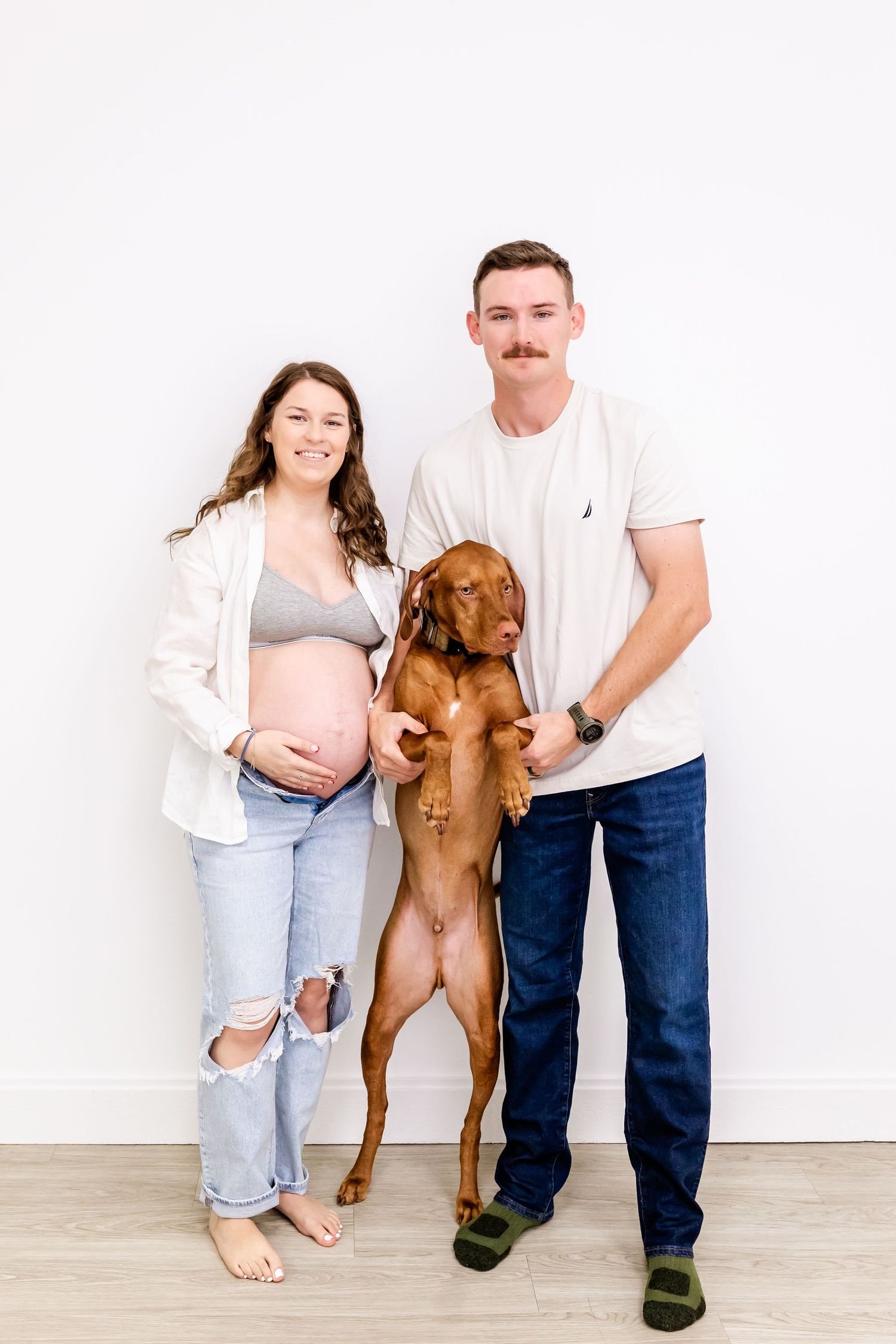 A couple in casual attire poses with their vizsla dog standing on its hind legs against a white wall.
