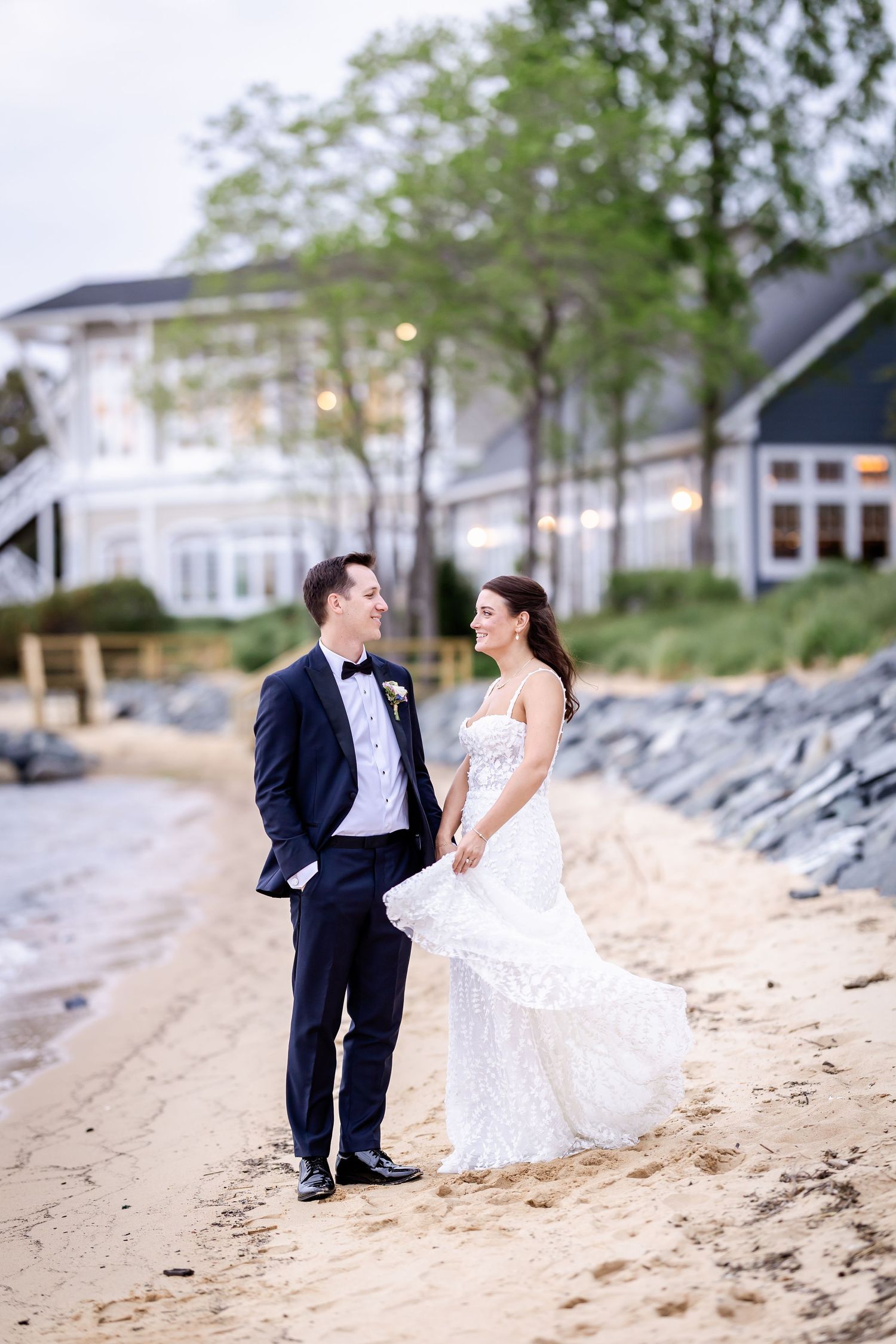 A couple poses romantically on the beach in formal attire with a coastal home behind them.