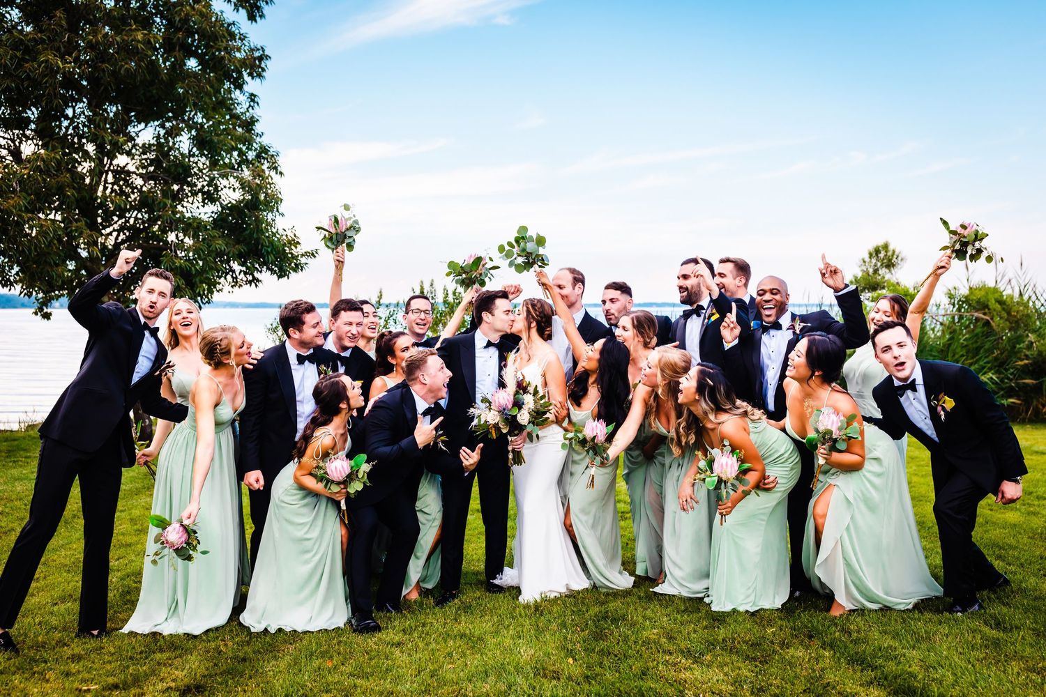 Wedding party in formal attire poses together on a lush green lawn during outdoor celebration.