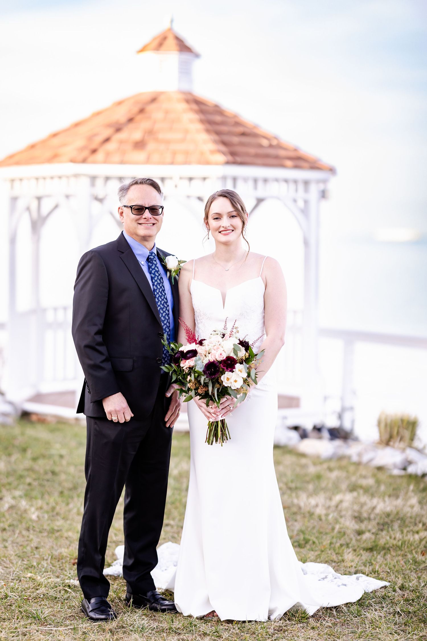 A wedding couple poses outdoors near a white gazebo on a grassy hill during their special day.