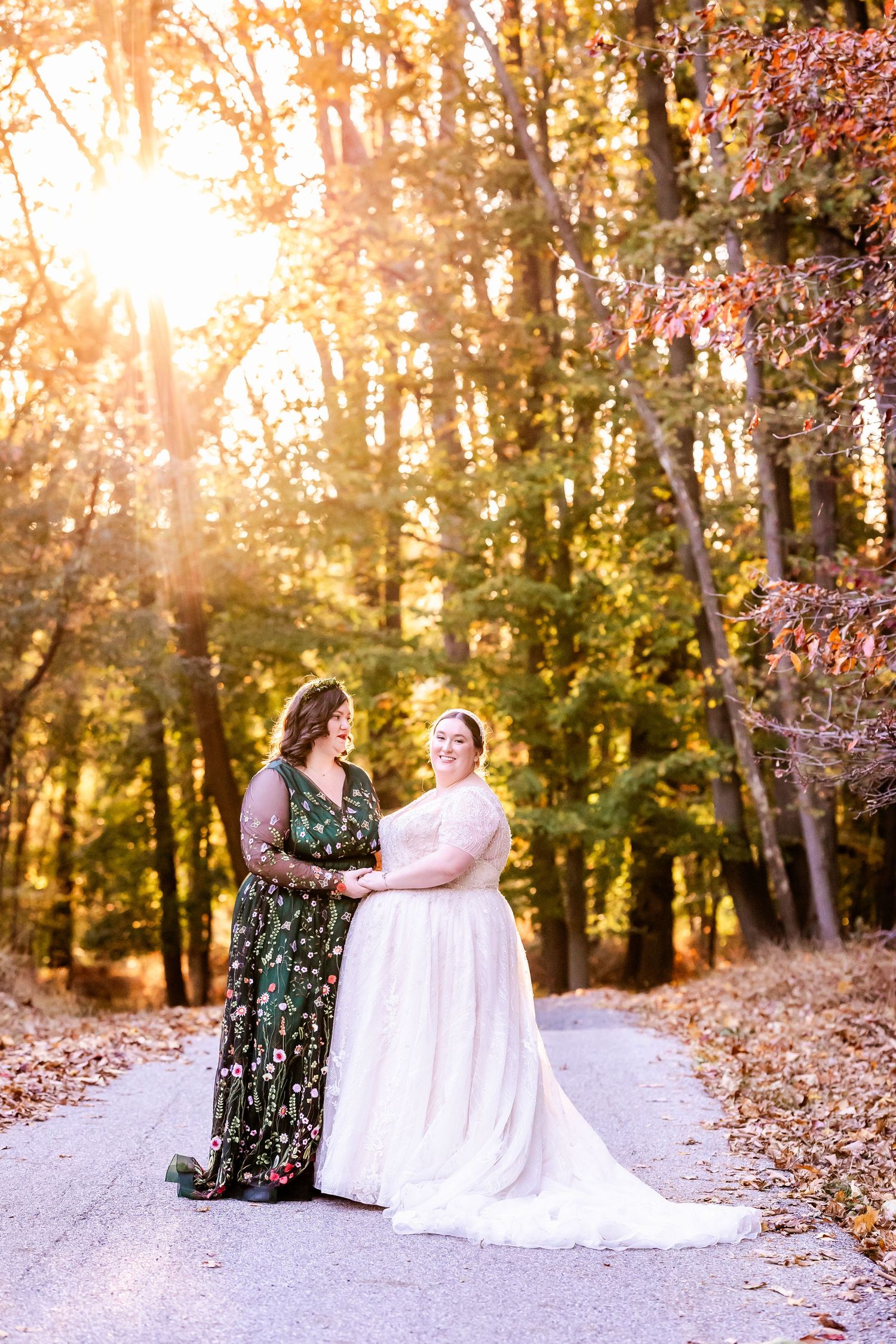 A romantic sunset wedding photo of two people in formal attire standing on a forest path with golden light streaming through trees.