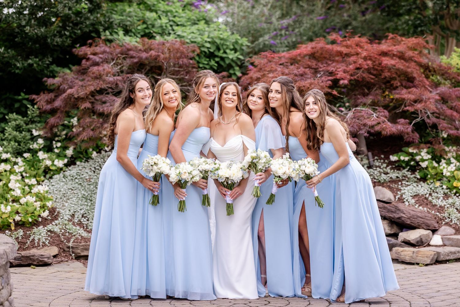 A group in light blue bridesmaid dresses and white bouquets pose together in a garden setting.