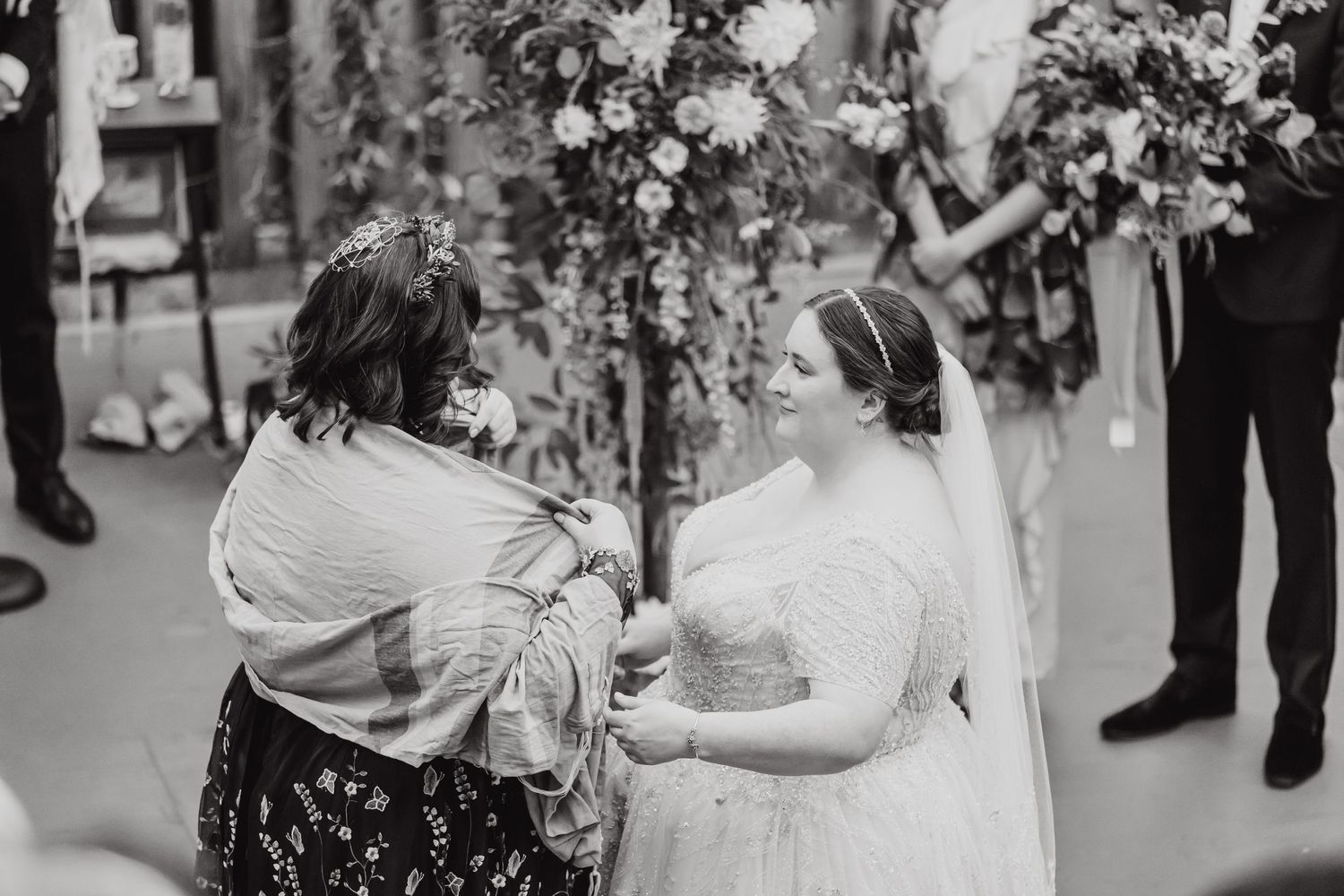 Black and white candid wedding moment featuring heartfelt interaction by floral arch.