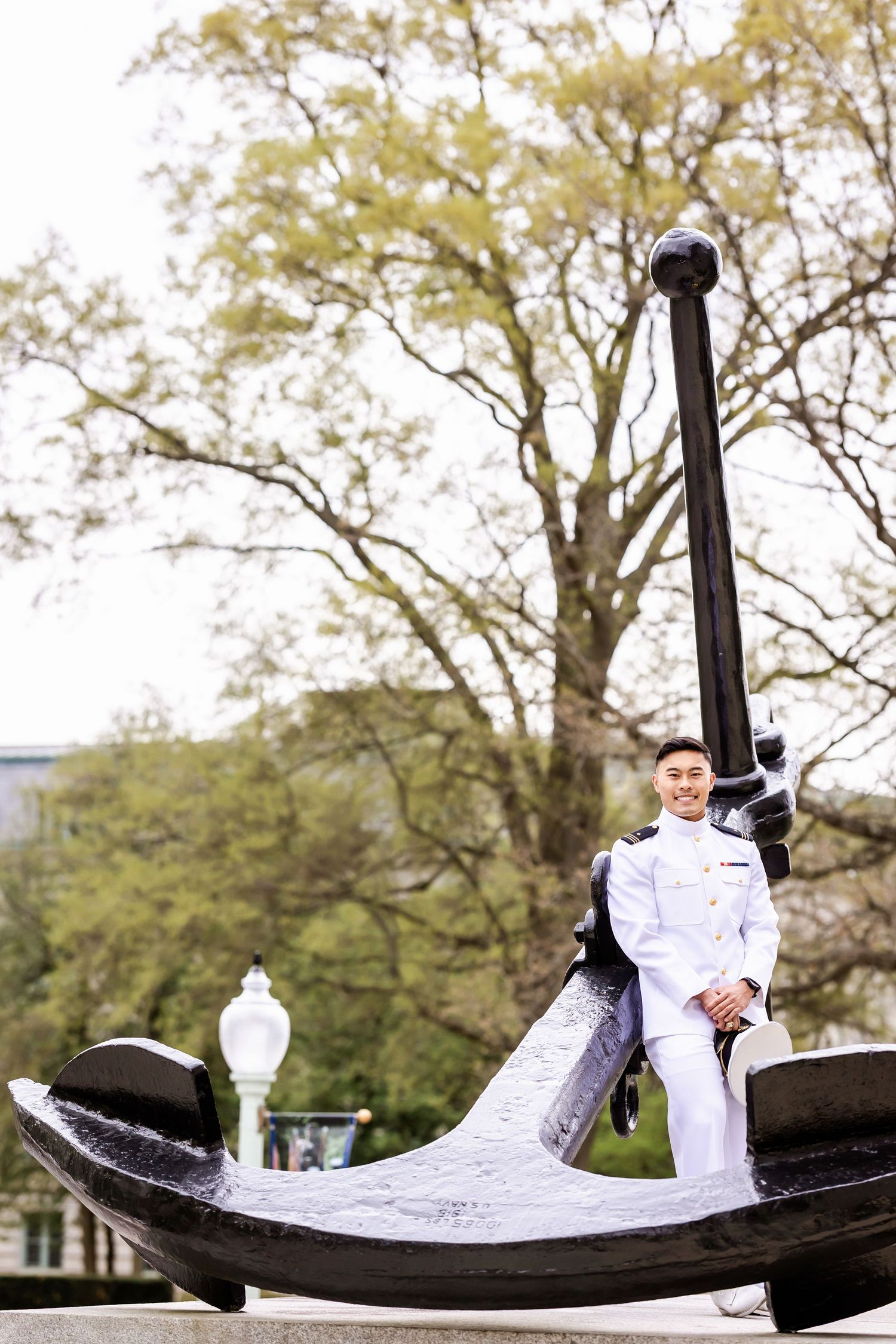 Naval officer in dress whites poses on a large decorative anchor monument in an outdoor park setting with autumn trees.