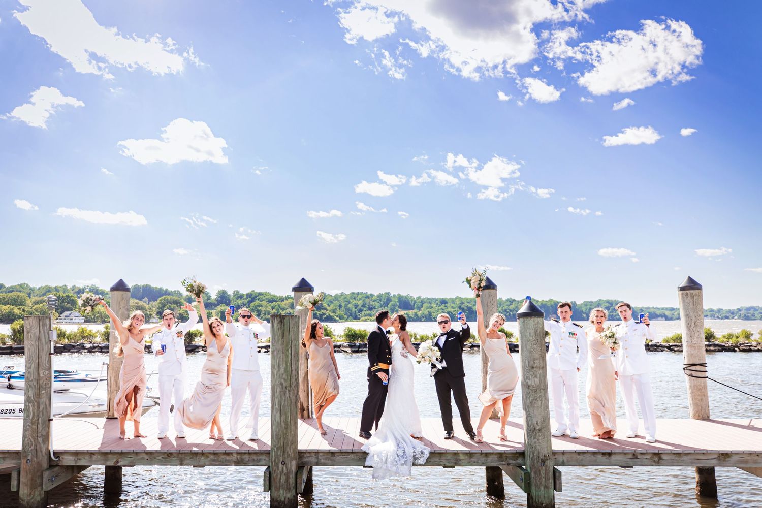 Wedding party stands on wooden dock overlooking waterfront under blue sky with white clouds.