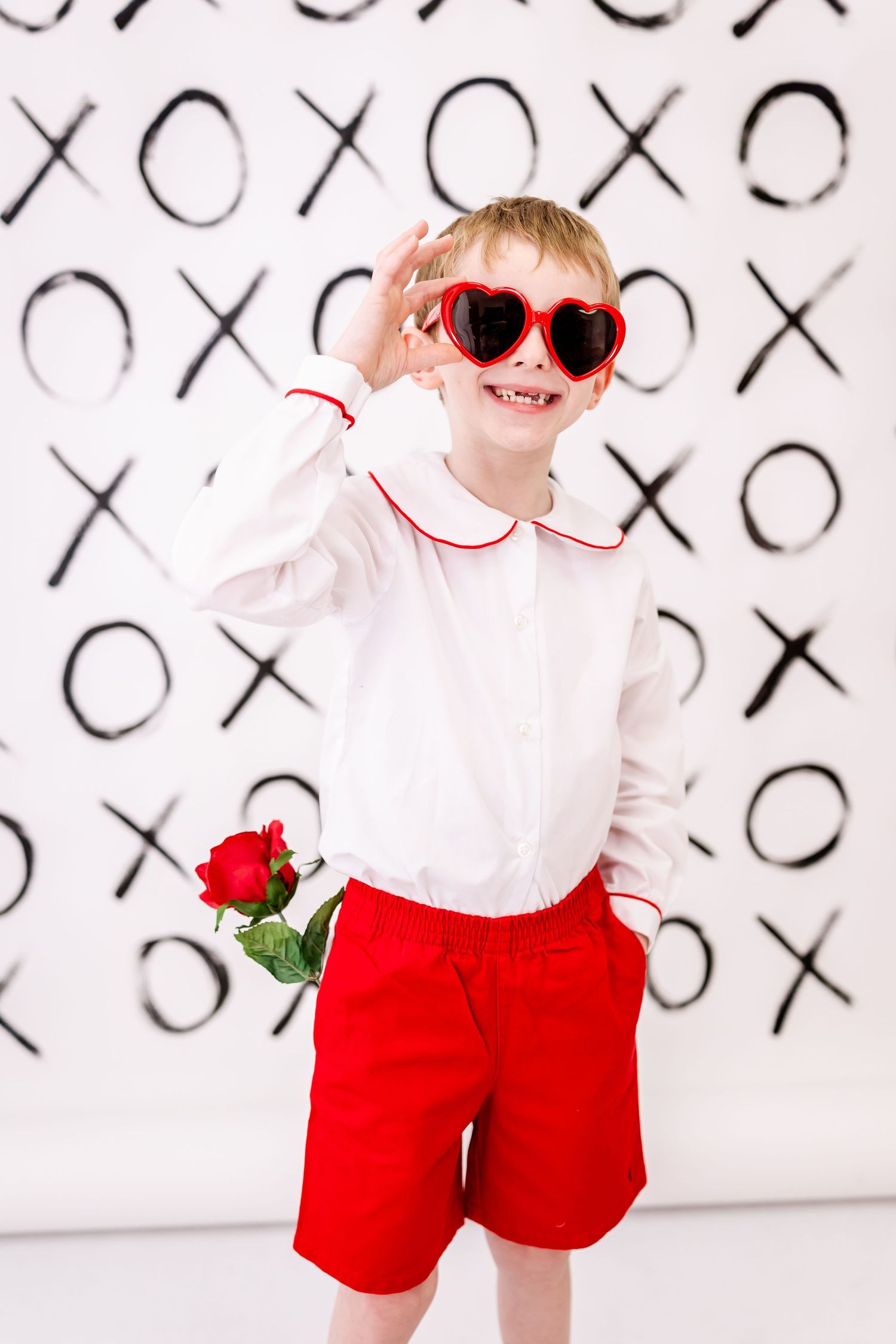 Young child in white shirt and red shorts poses with heart sunglasses against a wall decorated with X's and O's.