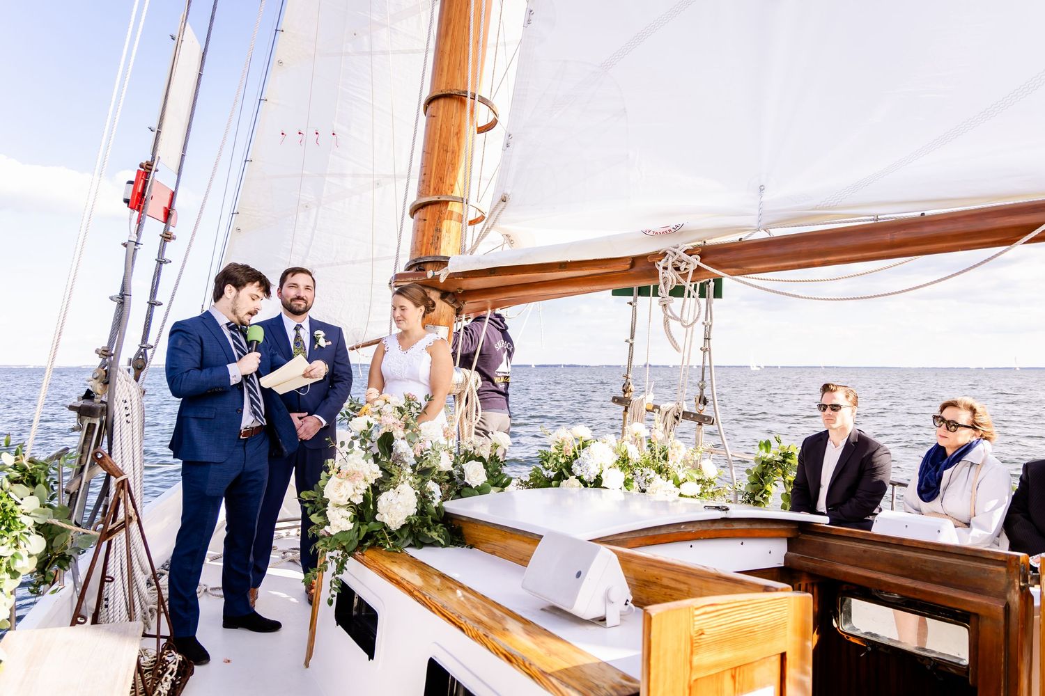 Couple exchanging vows on a decorated sailboat wedding ceremony with floral arrangements and guests on open water.
