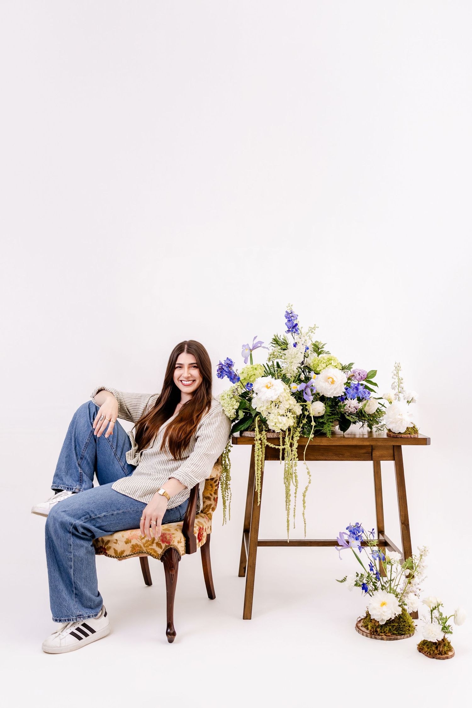 Smiling woman in jeans sits beside a wooden table with blue and white floral arrangements on a white background.