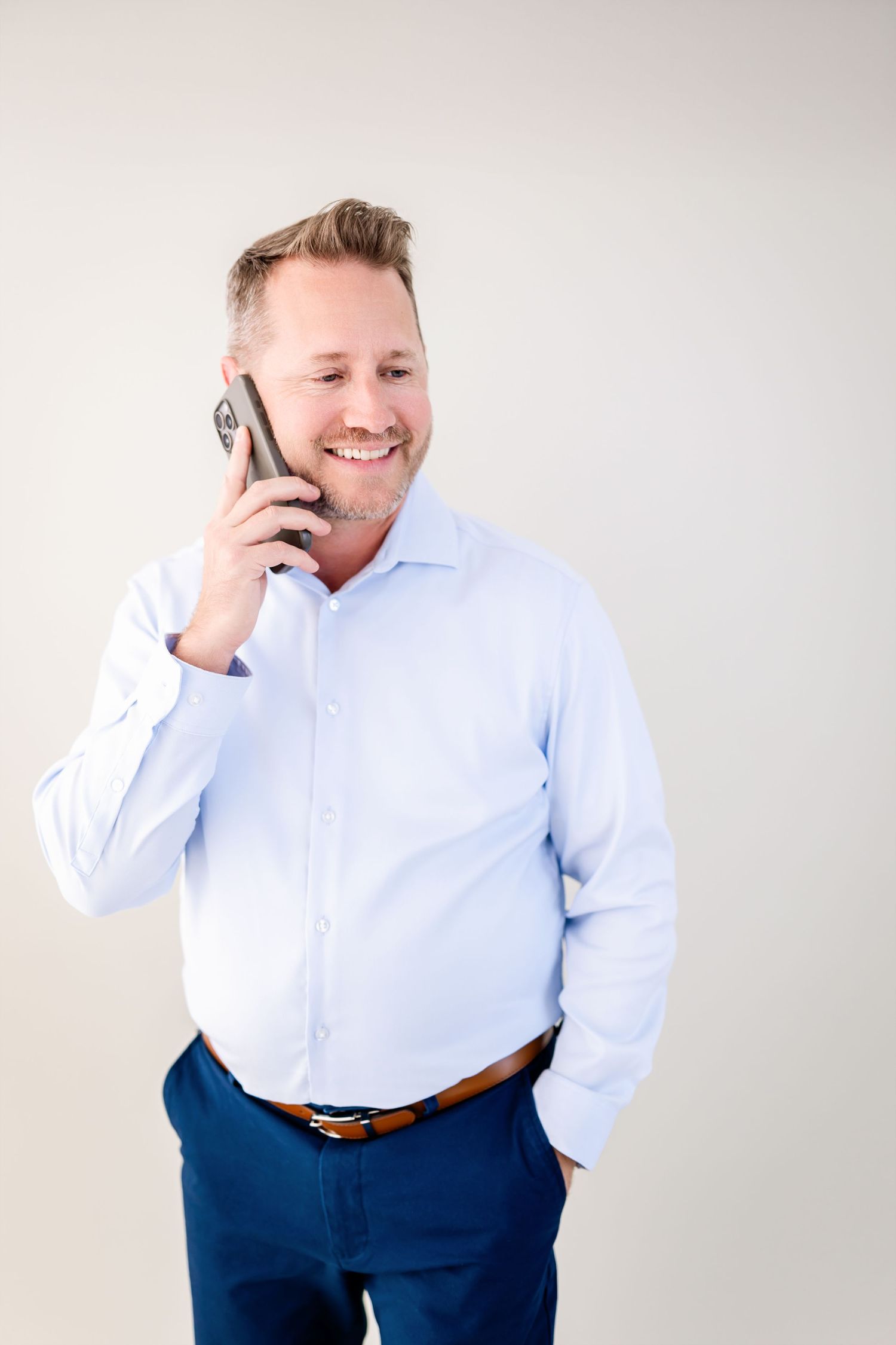 Smiling businessman in light blue shirt and navy pants talking on smartphone against white background.