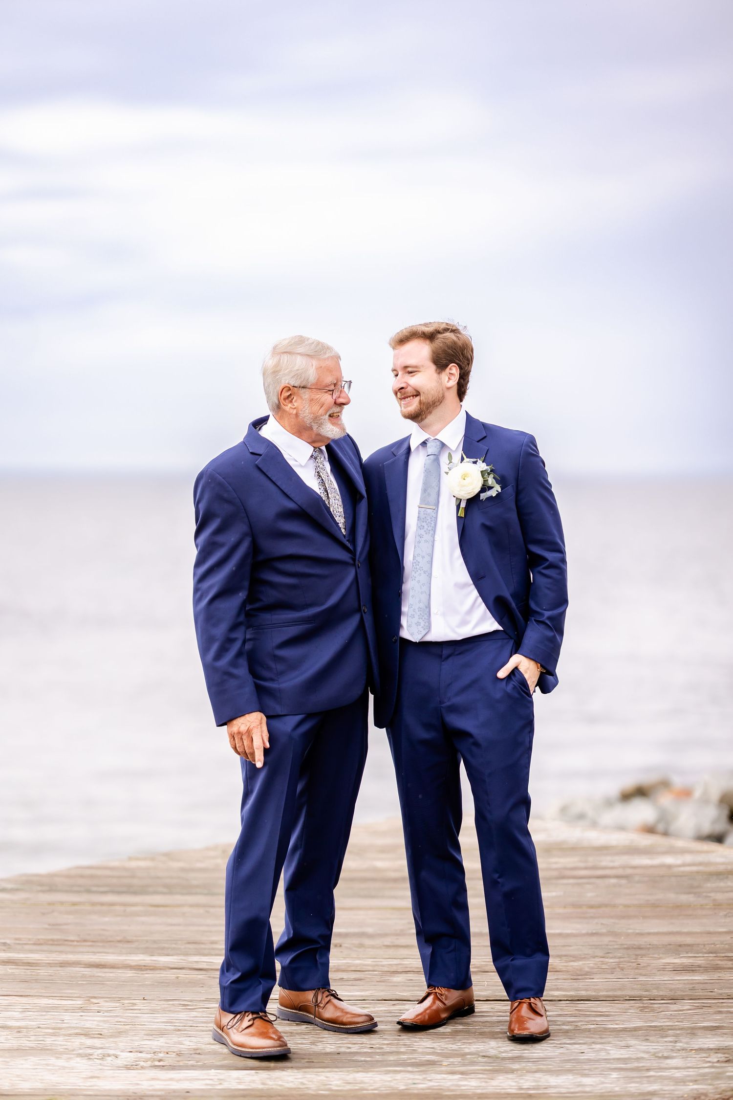 Two people in navy suits share a lighthearted moment on a wooden boardwalk by the ocean.