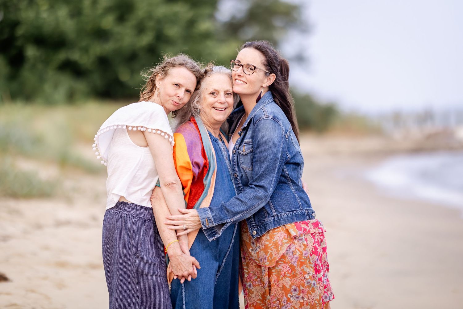 Three friends share a joyful moment together while standing on a beach with coastline in the background.