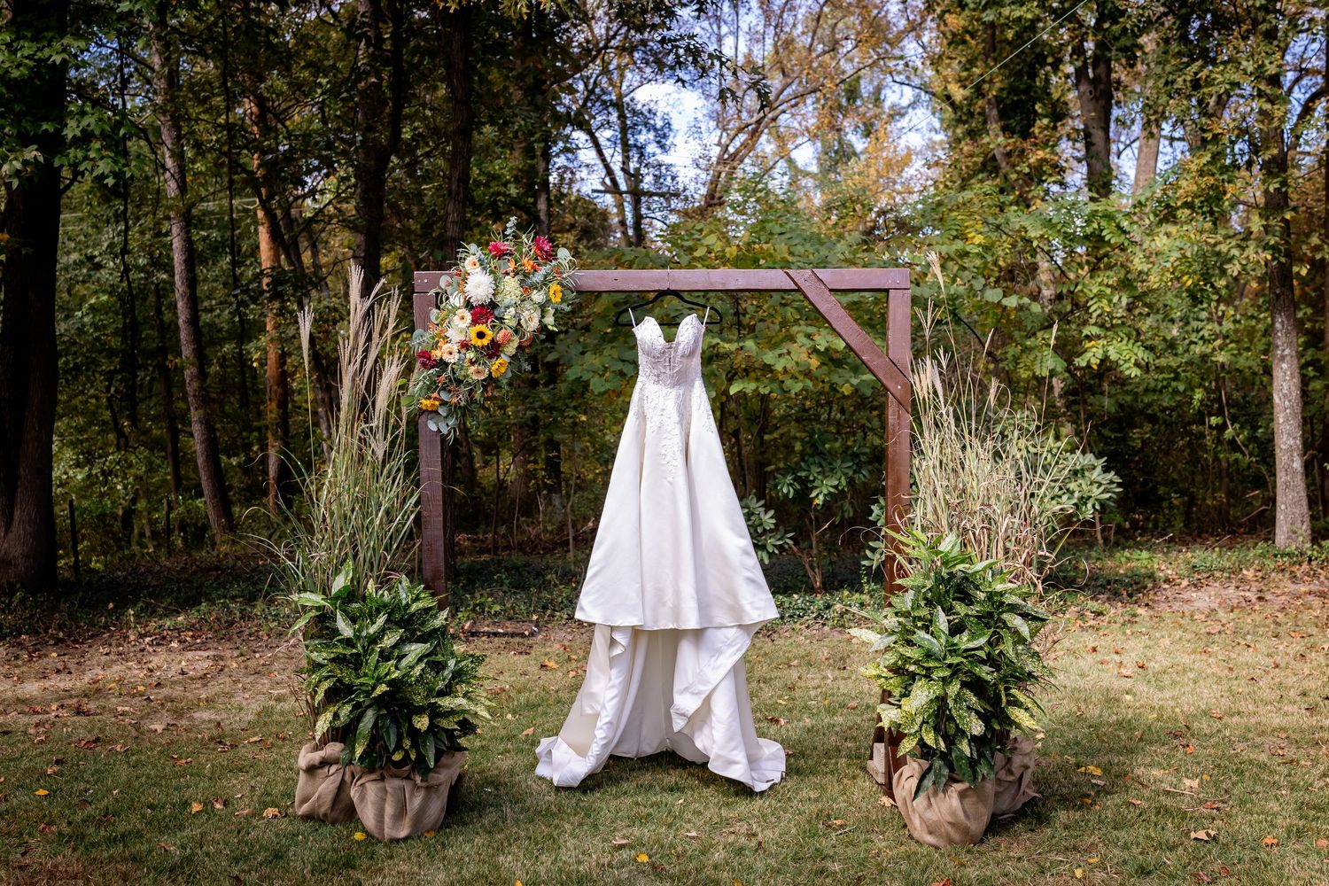 A white wedding dress hangs on a rustic wooden arch in a forest setting with decorative potted plants at the base.