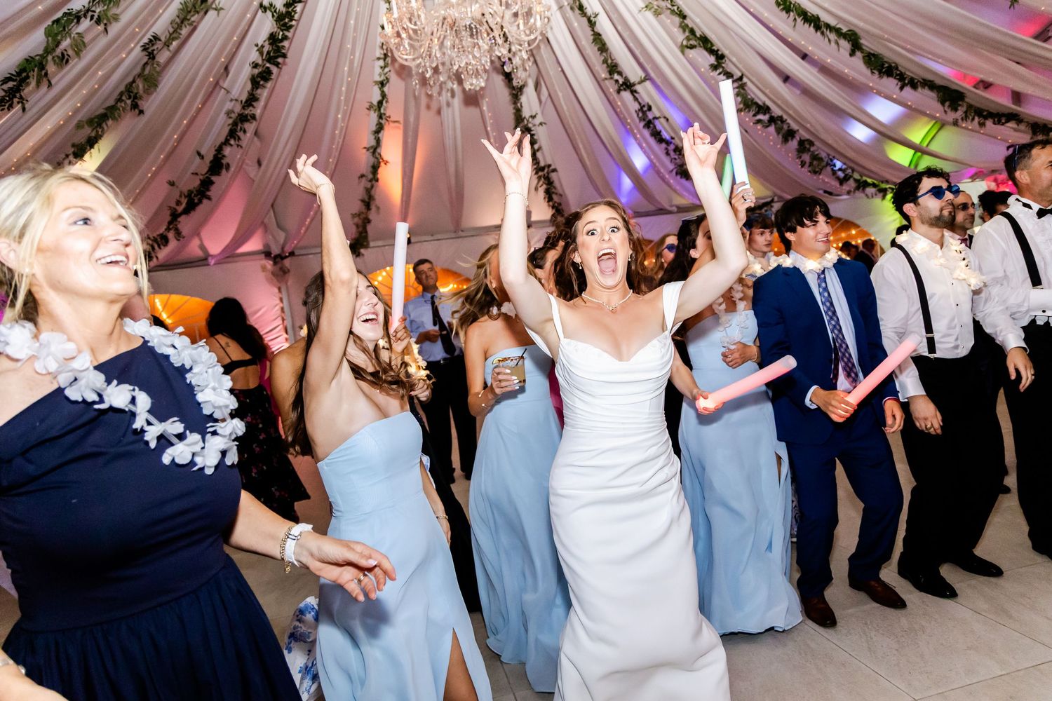 Wedding guests dance together in a reception hall decorated with pink lighting and elegant white drapery.