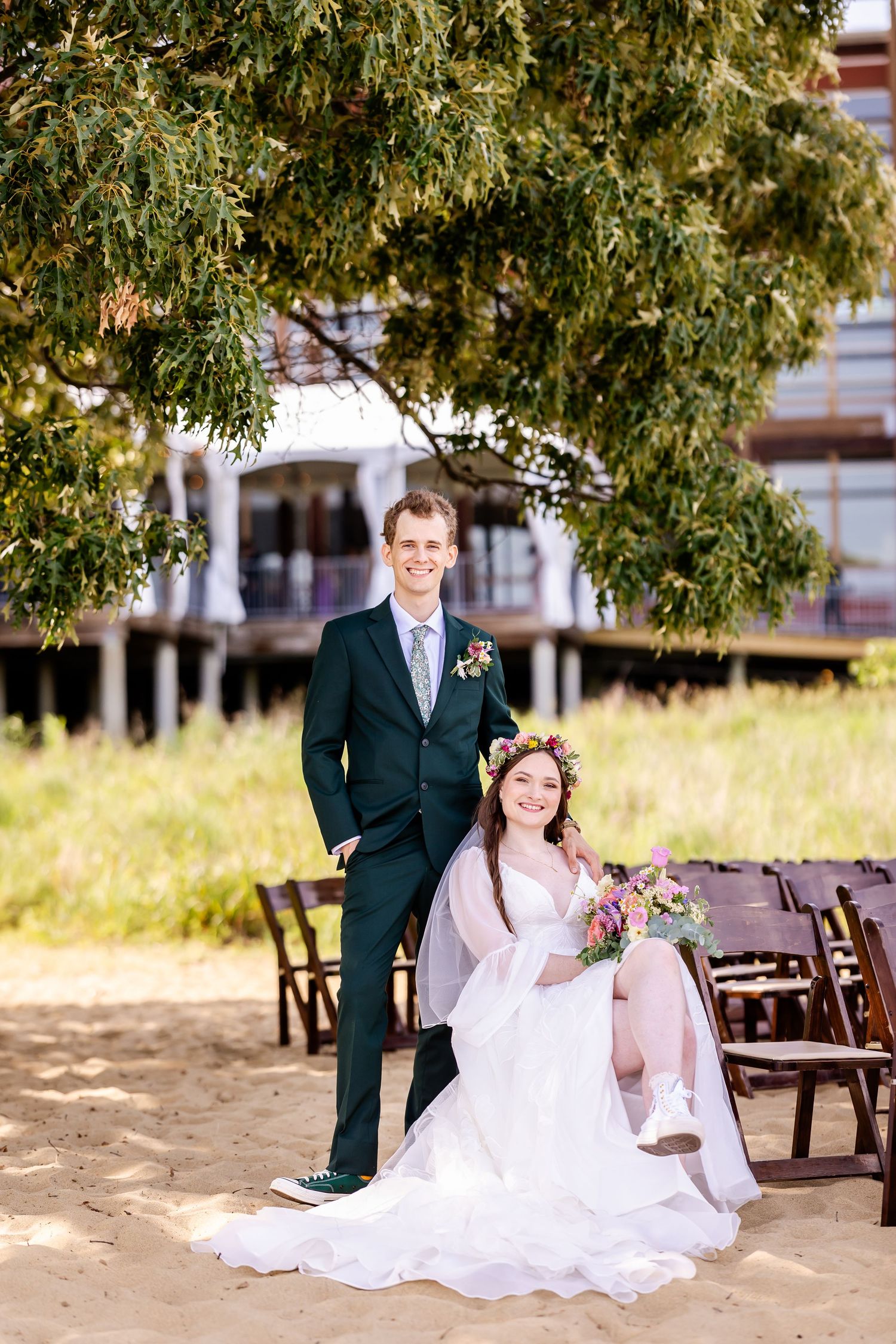 A couple in formal attire poses together under a tree on a sandy beach with wooden benches in the background.