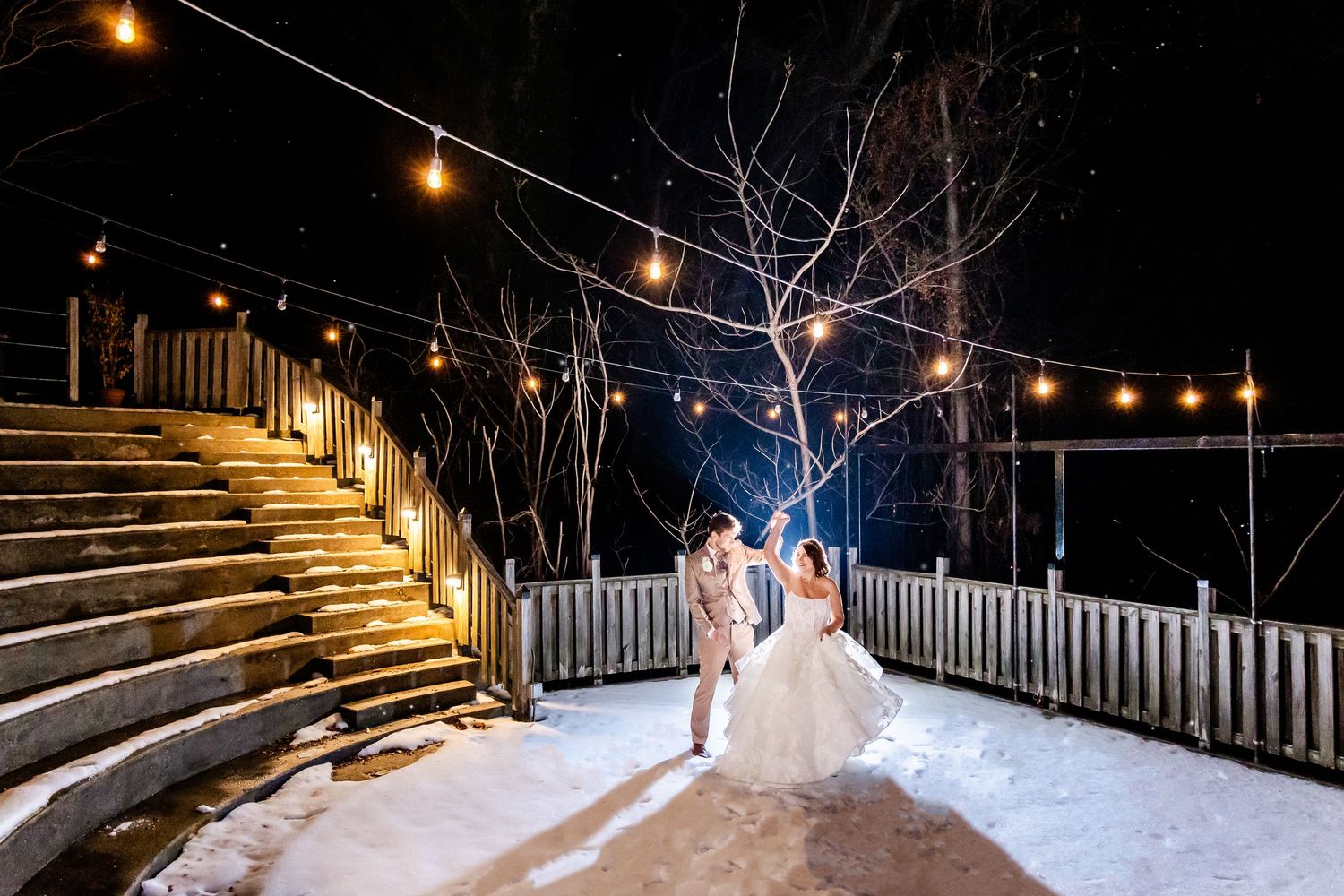 String lights illuminate a snowy outdoor deck with wooden stairs and railing during a winter night.