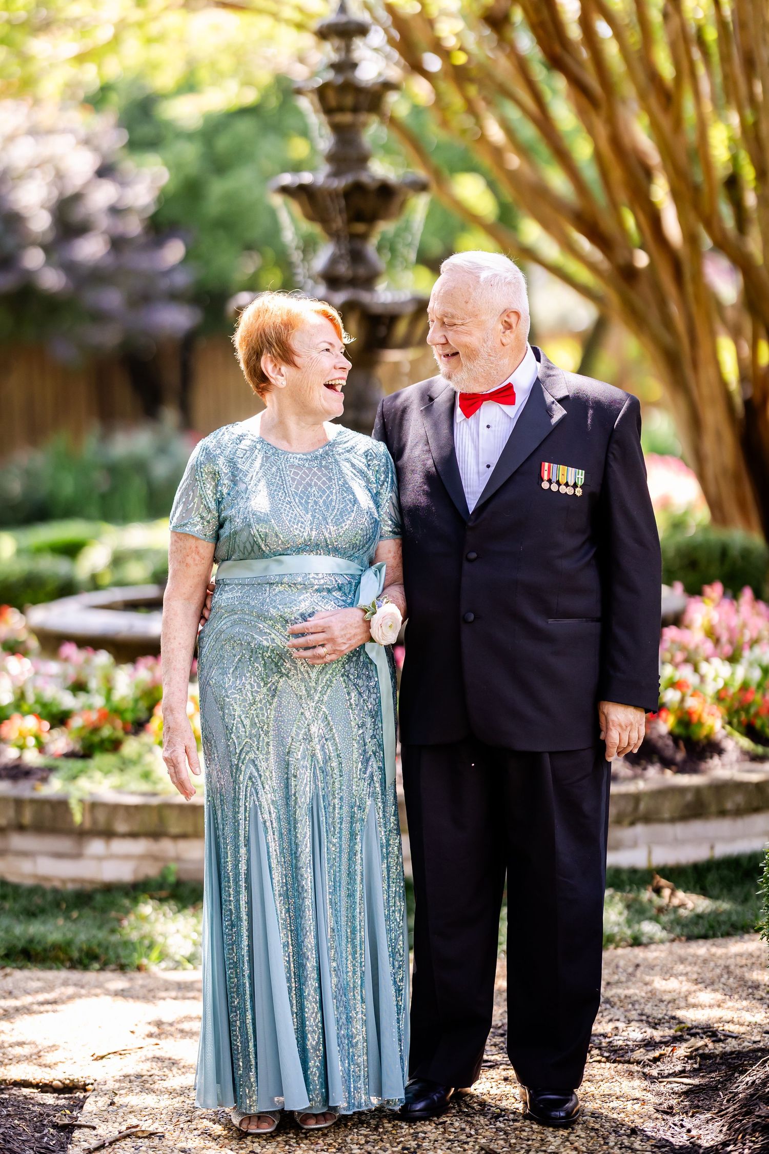 Couple in formal attire poses together in a garden setting with the woman wearing a sparkly blue dress.