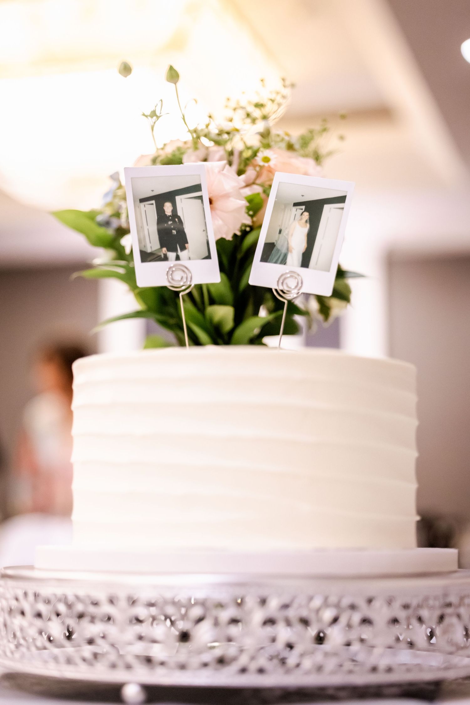 White wedding cake decorated with small floral accents and cake toppers on lace stand.