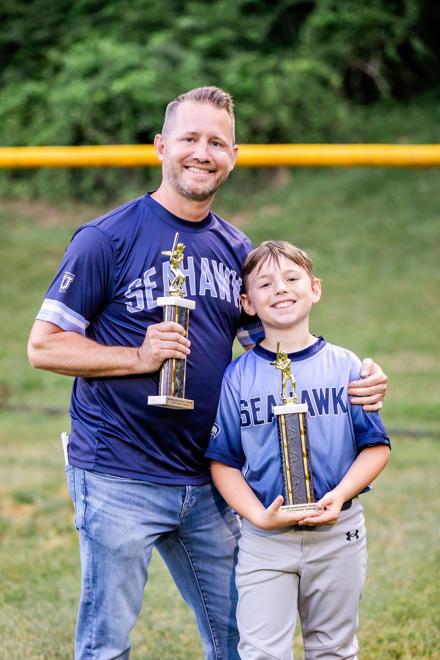 Father and son pose proudly with a baseball trophy on the field.