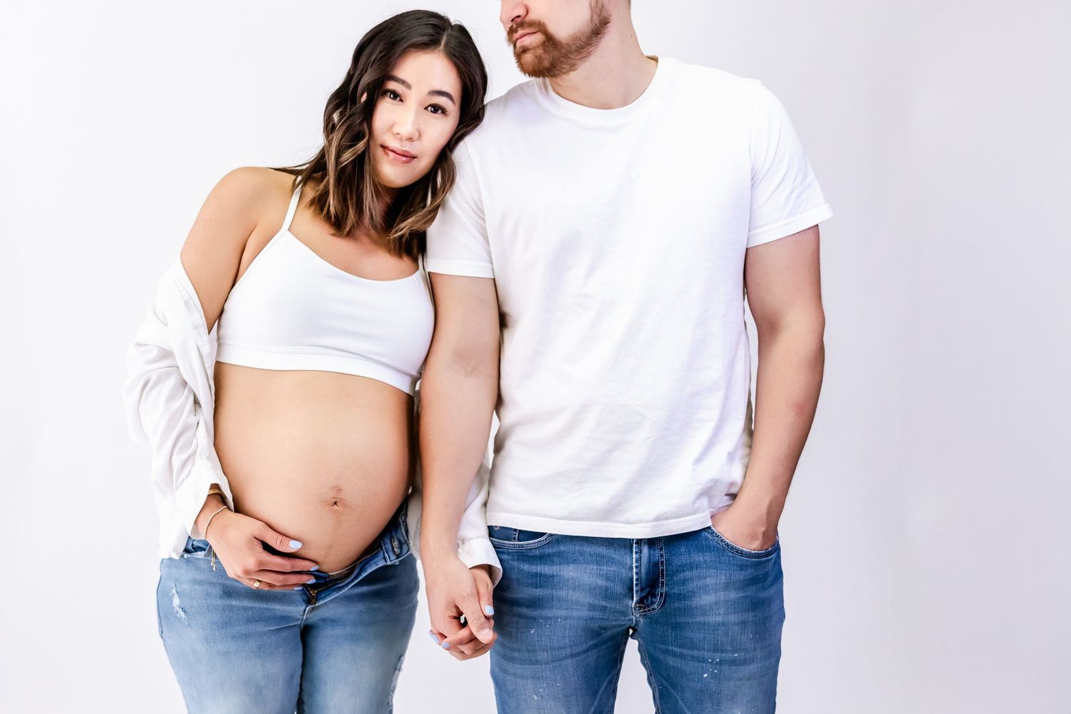 Minimalist maternity photo of couple in white and denim against plain background.
