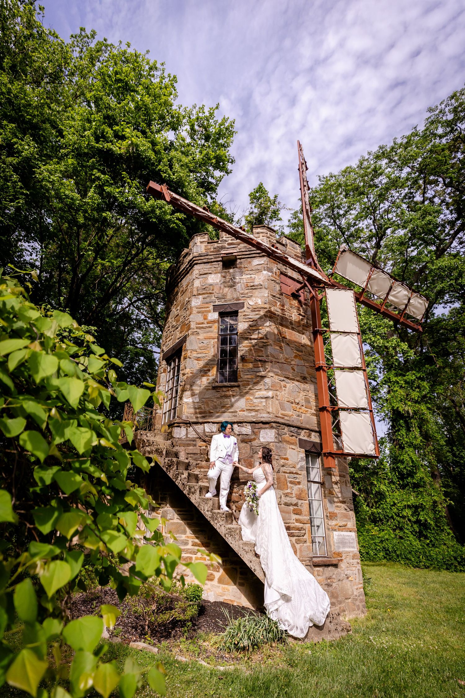 A wedding couple stands against an old stone tower structure with dramatic cloudy sky backdrop.