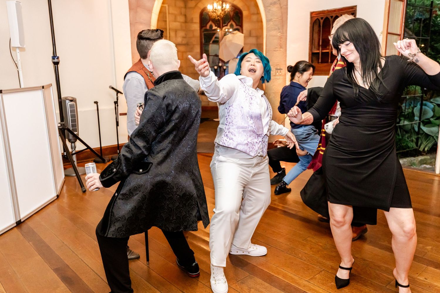 Party guests dancing enthusiastically at indoor celebration with wood floors and stone archways.