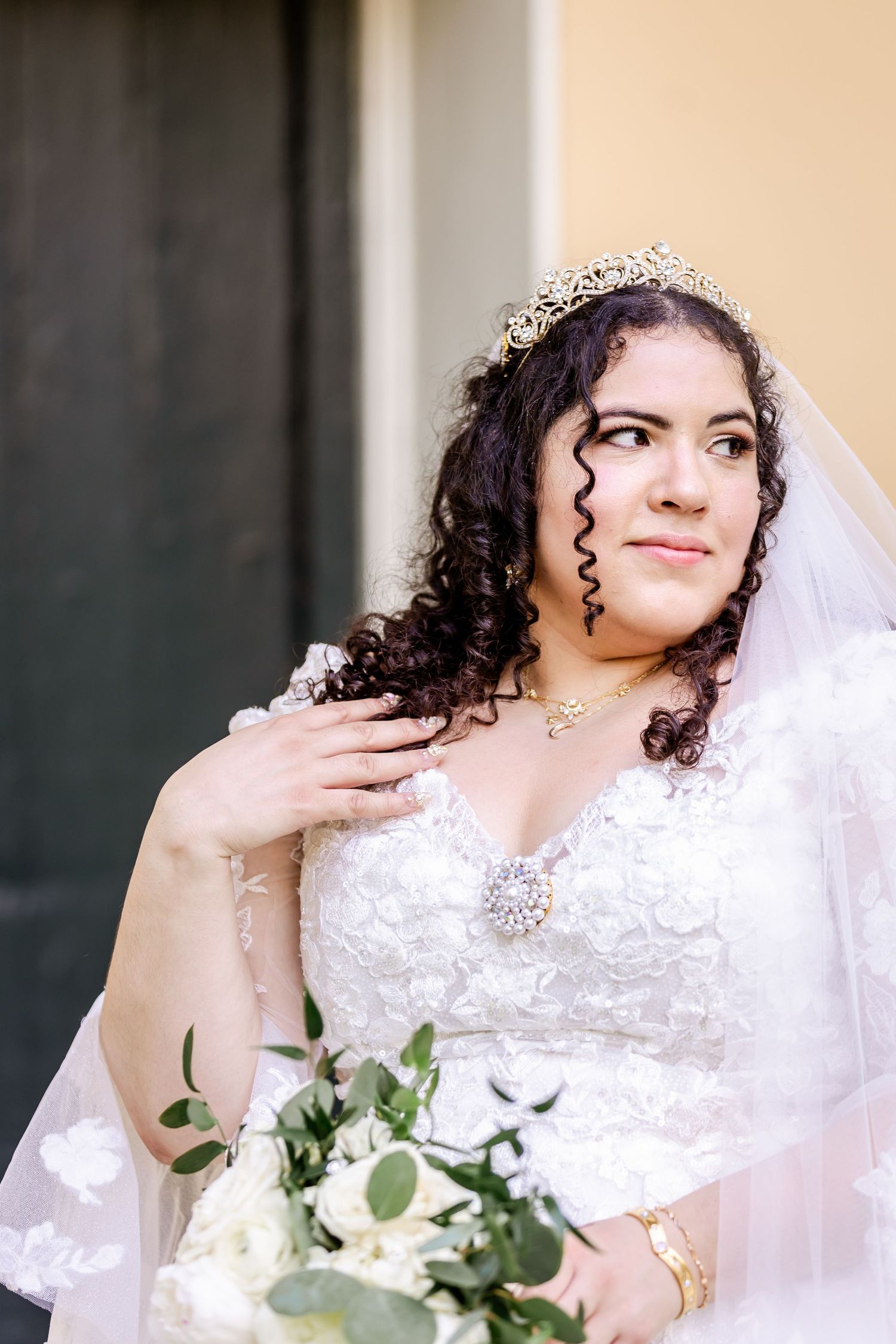 Classic bridal portrait featuring a delicate tiara, flowing veil, and white floral bouquet against a neutral wall.