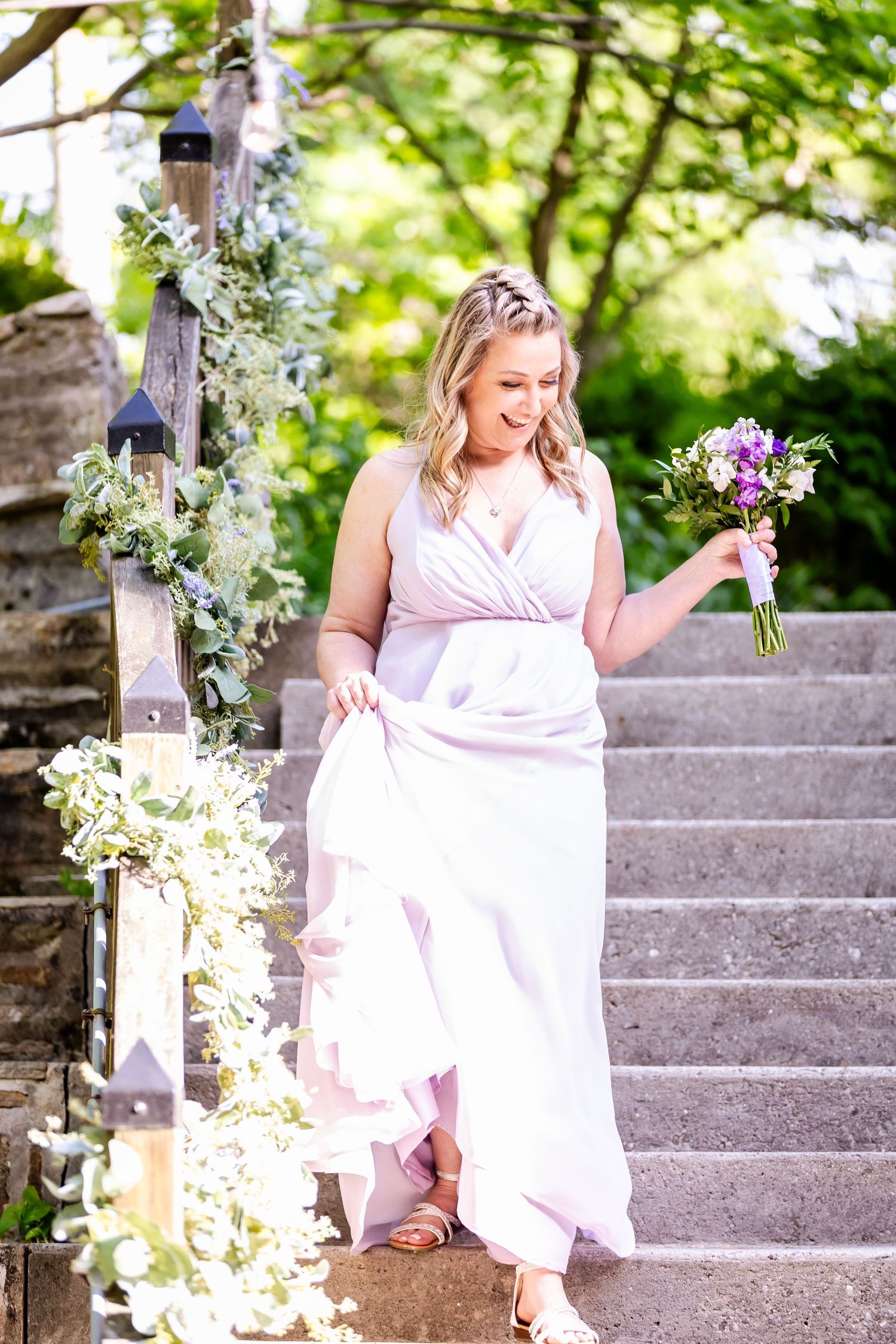 Woman in flowing white dress holds bouquet while standing on decorated outdoor stone staircase.
