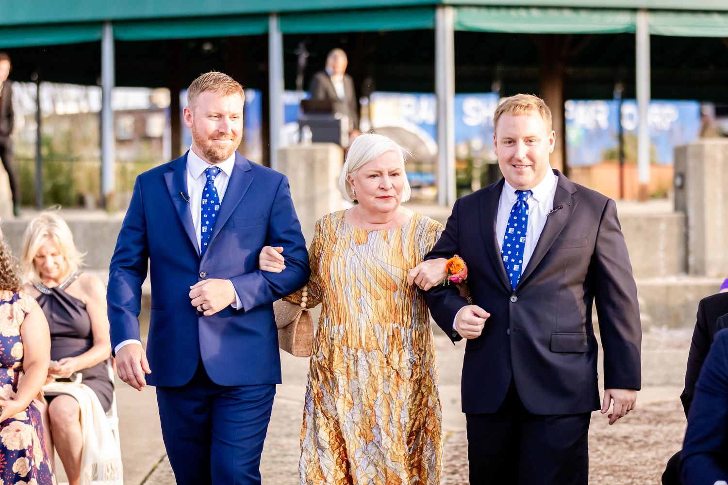 Three people in formal attire walk together on a sunny day, with a woman in gold between two men in blue suits.