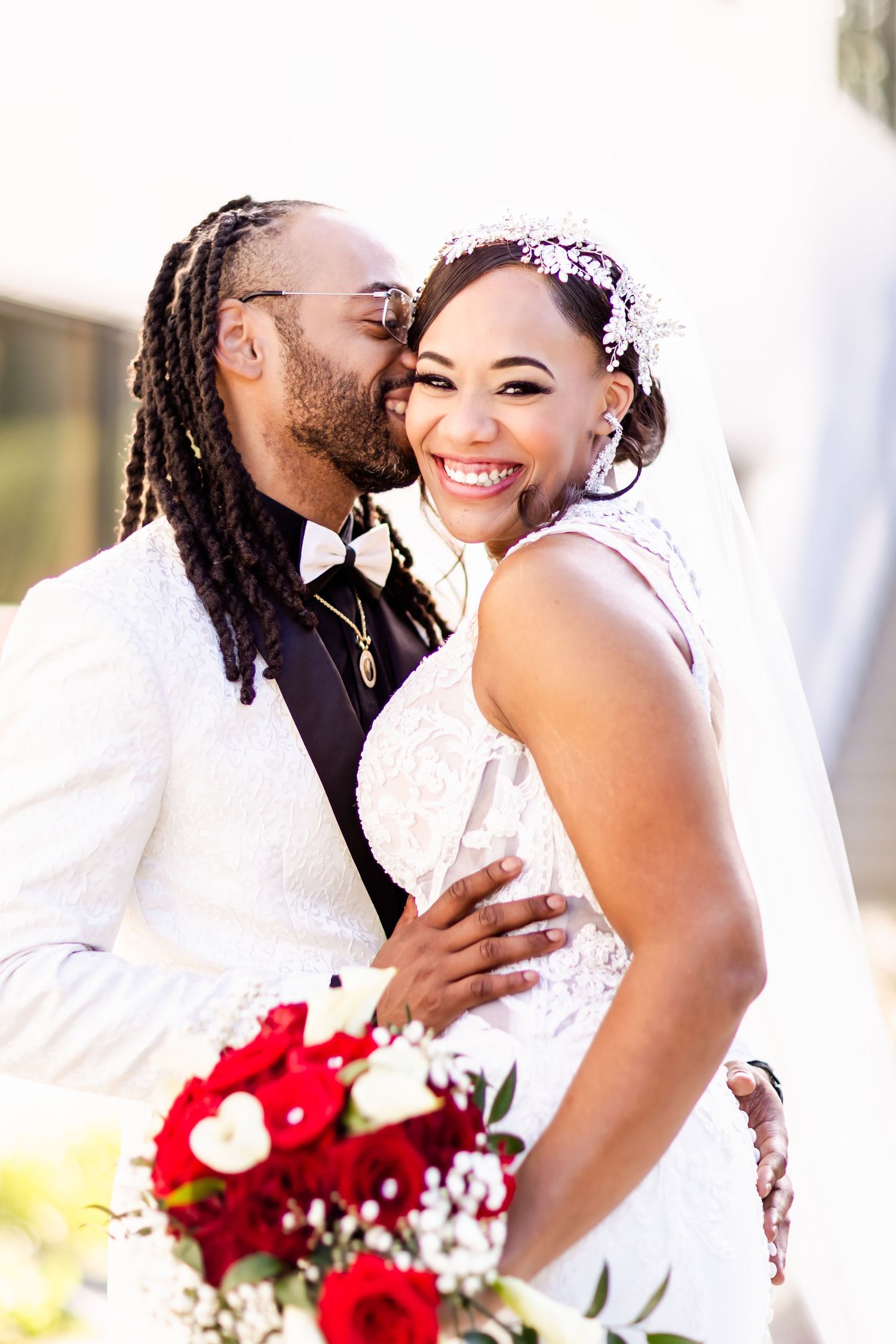 Joyful couple embracing during wedding photoshoot with vibrant red floral bouquet.