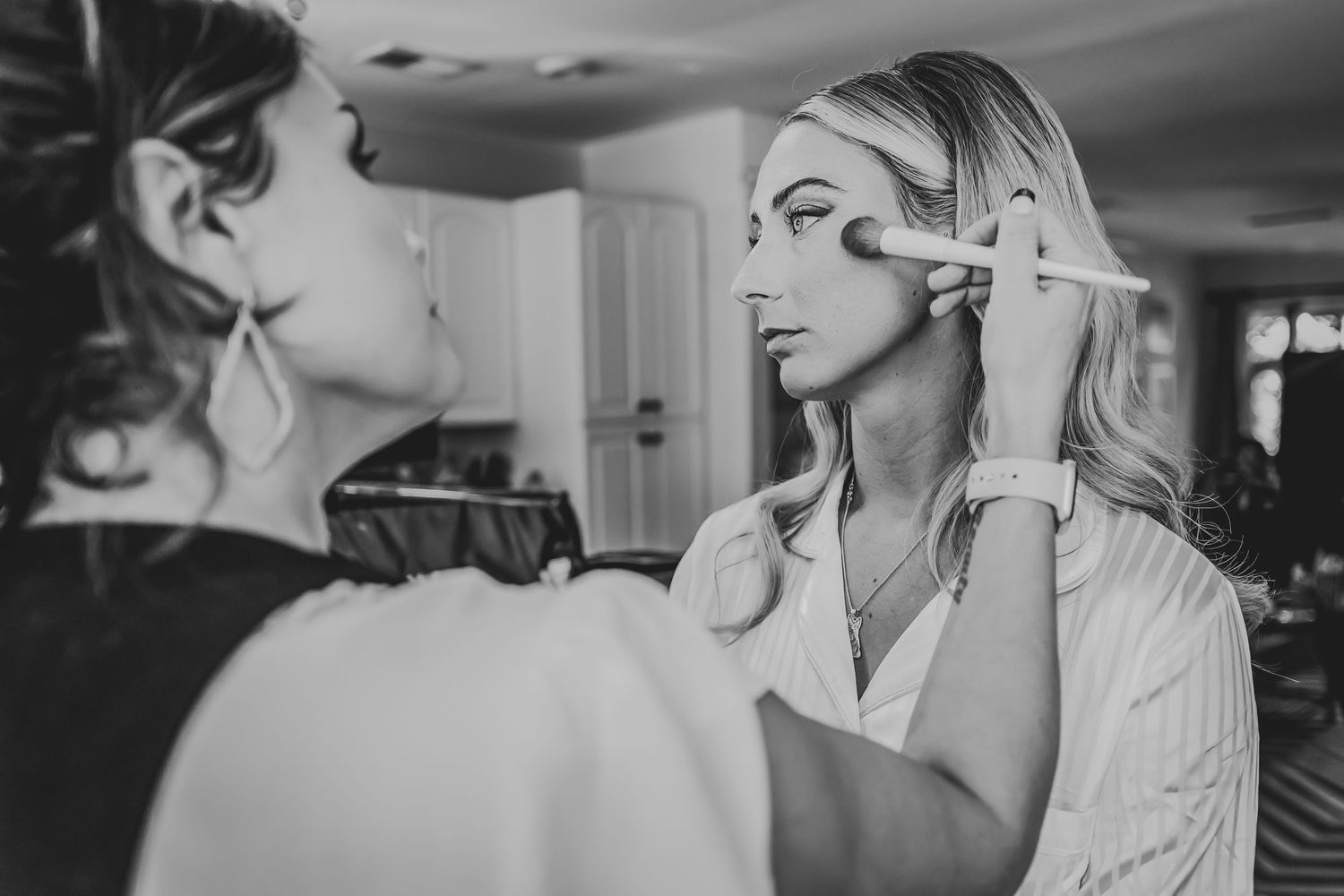 A black and white image showing makeup application in progress as a stylist works with a client in a bright room.