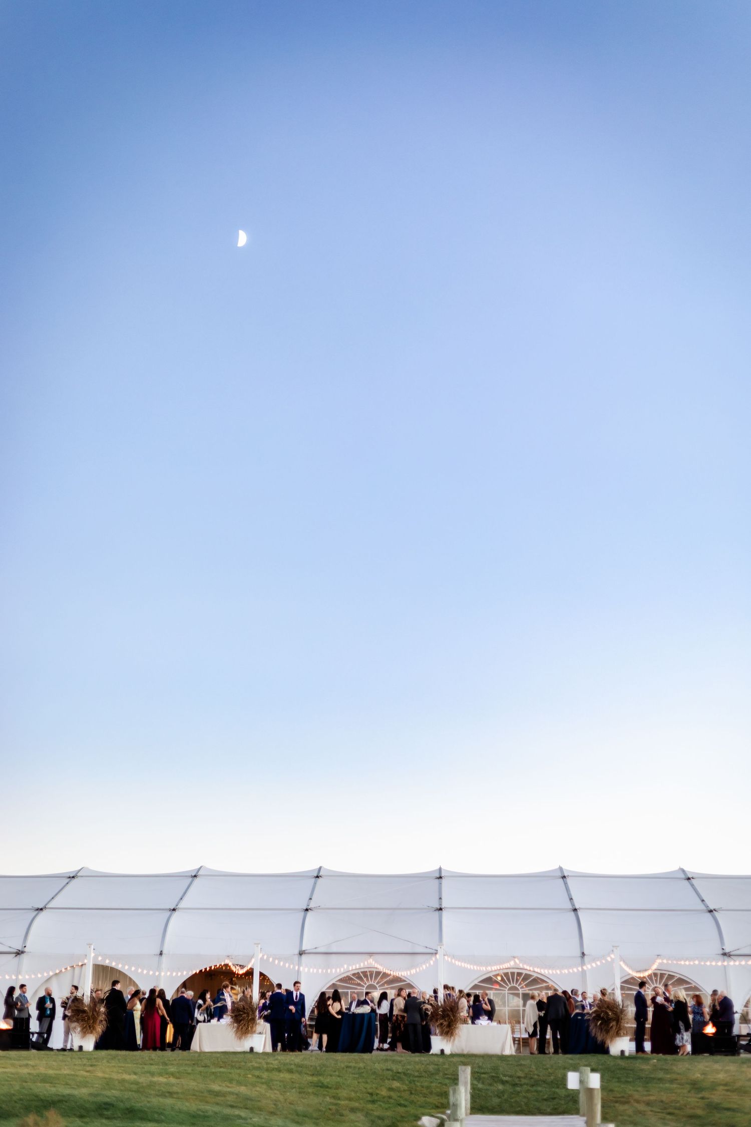 Large white wedding tent setup against blue sky with moon visible during sunset.
