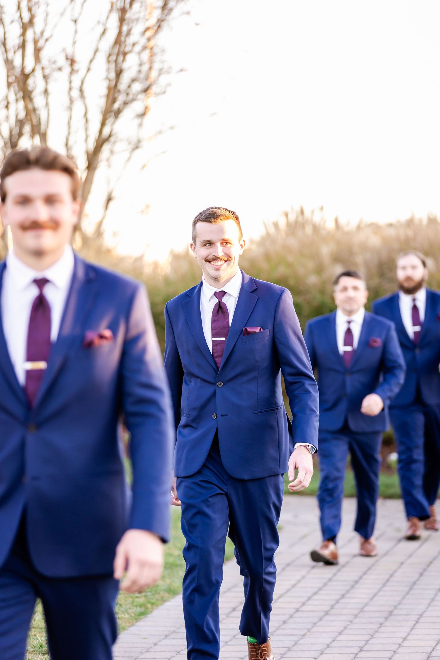Groomsmen in matching navy suits and burgundy ties walk along a garden pathway.