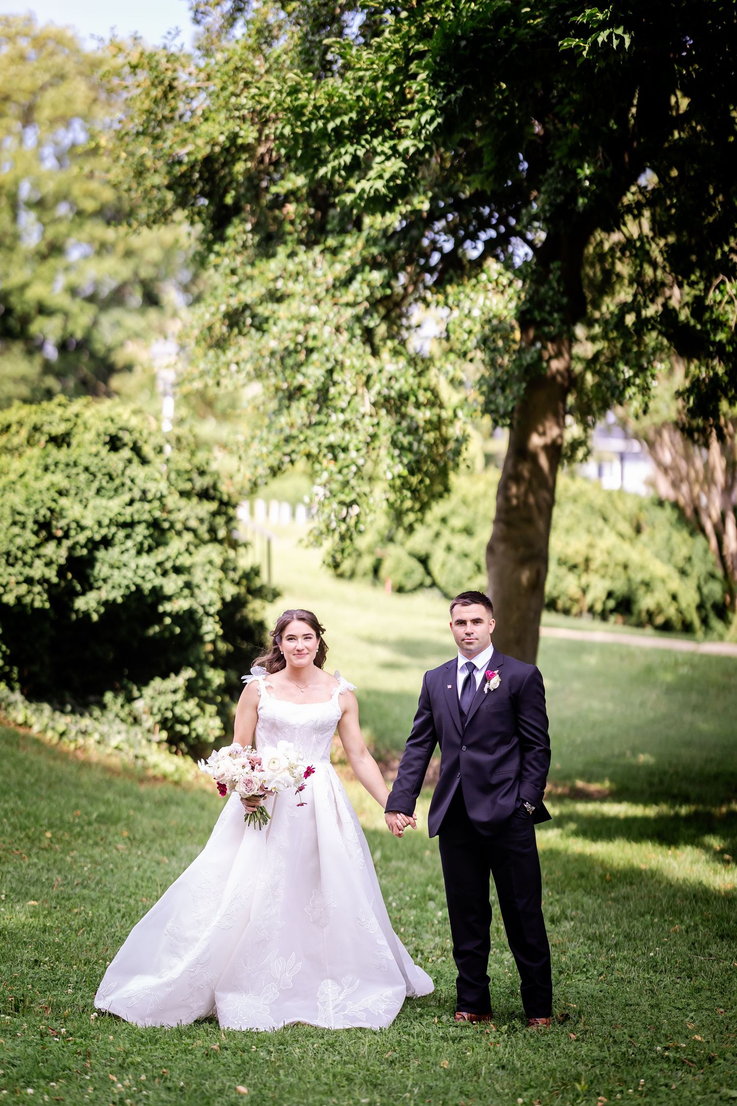 A wedding couple poses for portraits on a lush green lawn under tall trees.