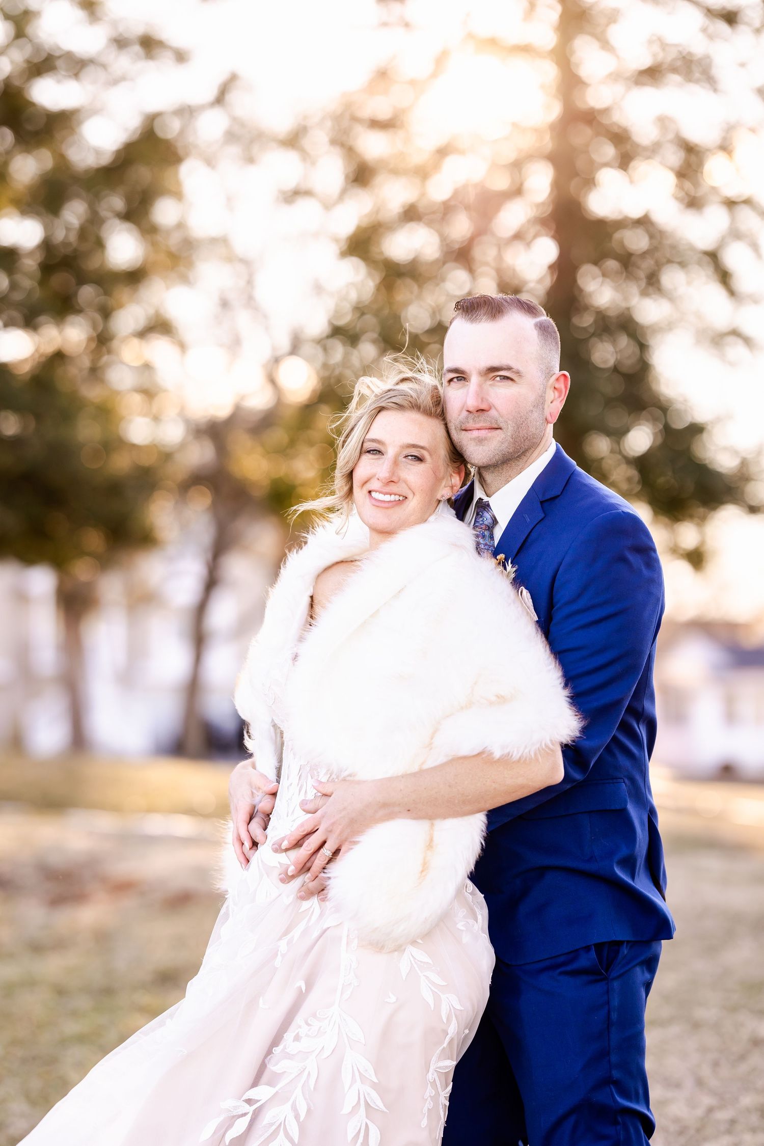 A joyful outdoor wedding portrait with the couple in blue suit and white gown with fur wrap.
