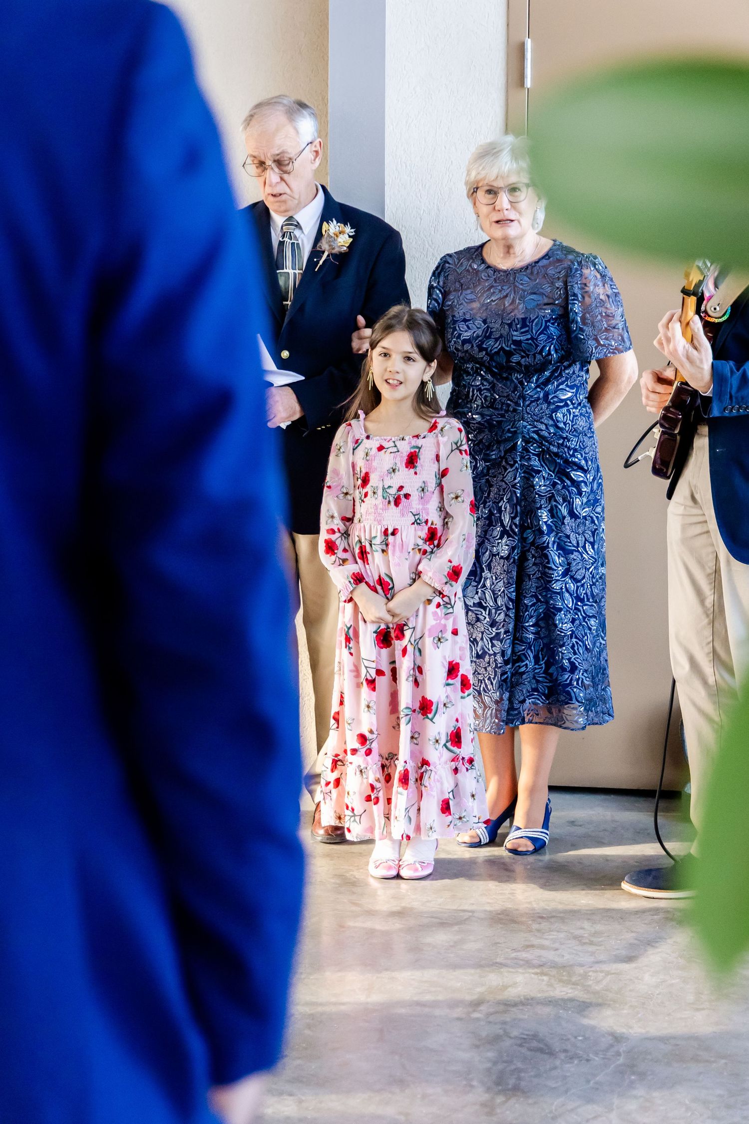 A young girl in a floral pink dress stands with two adults dressed formally during what appears to be a ceremony.
