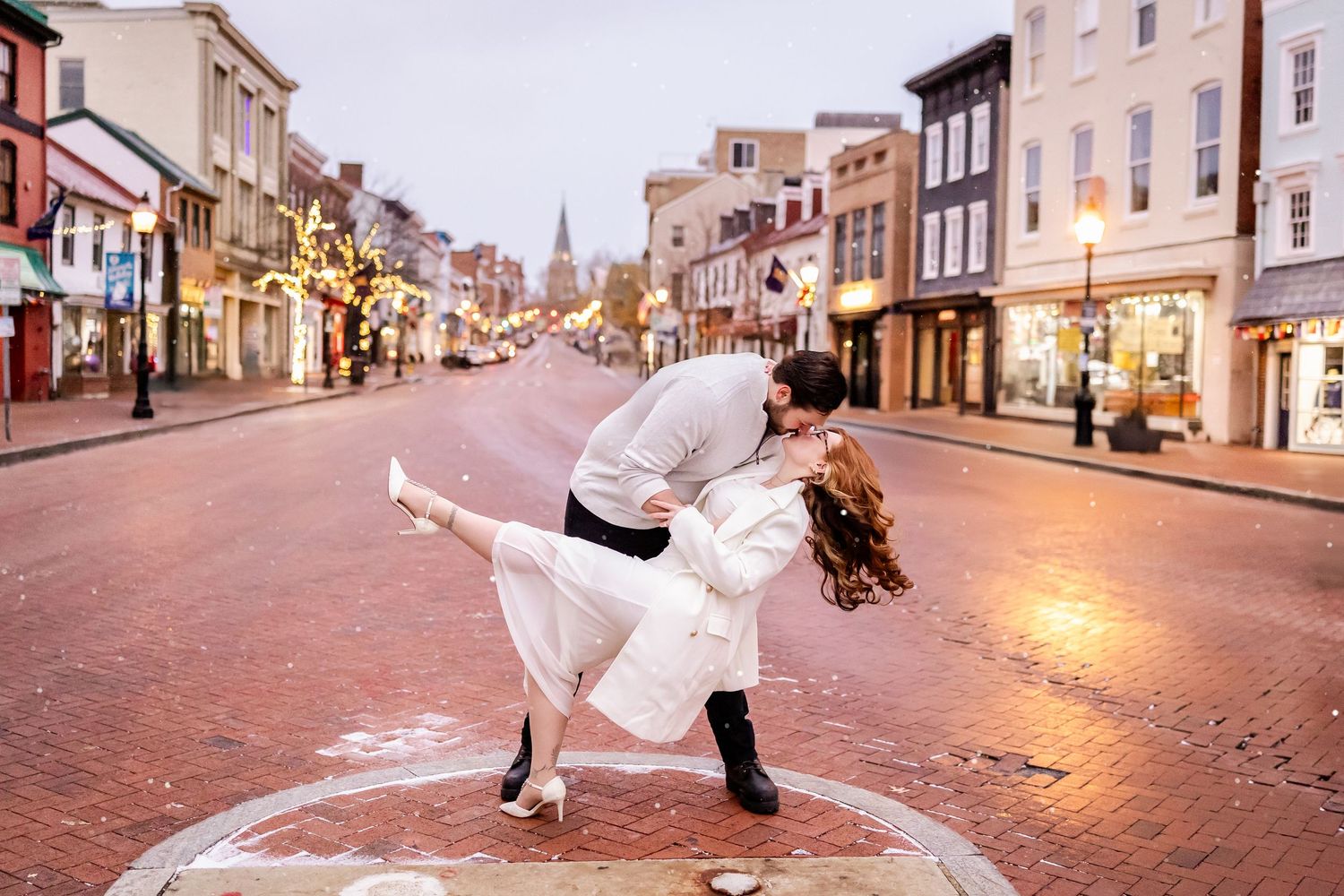 Couple in white winter outfits dancing together on a brick paved street during sunset in a charming downtown area.