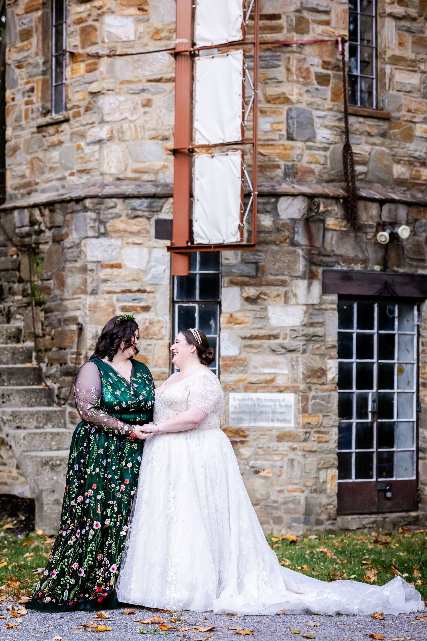 Two people in elegant dresses, one white and one green floral, stand together against a rustic stone building.