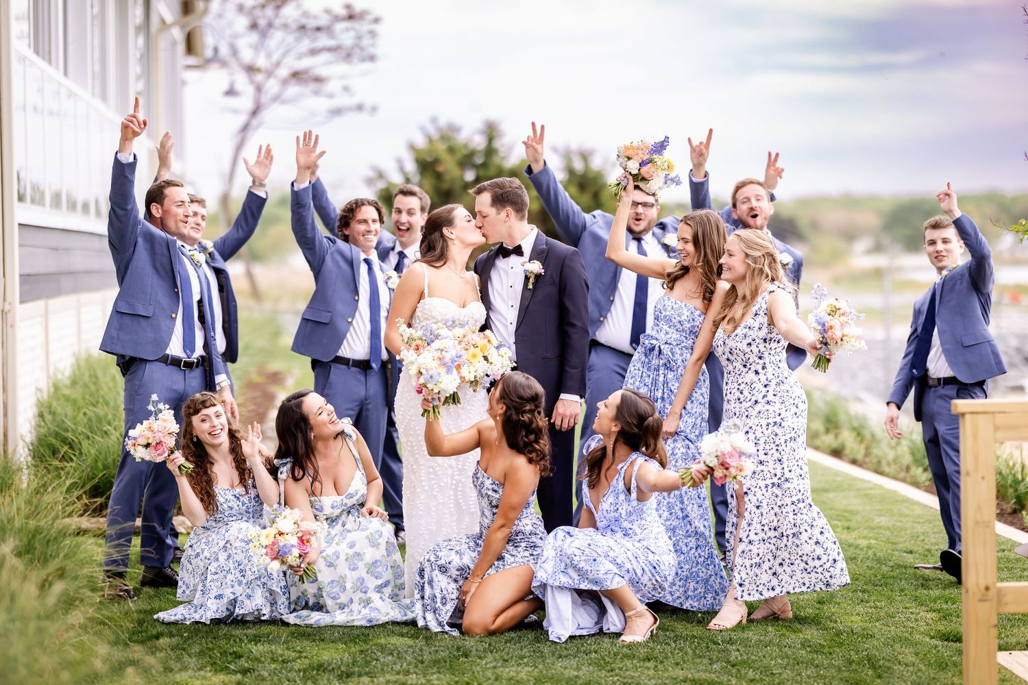 Wedding party celebrates with arms raised on sunny hillside in coordinated blue and floral attire.