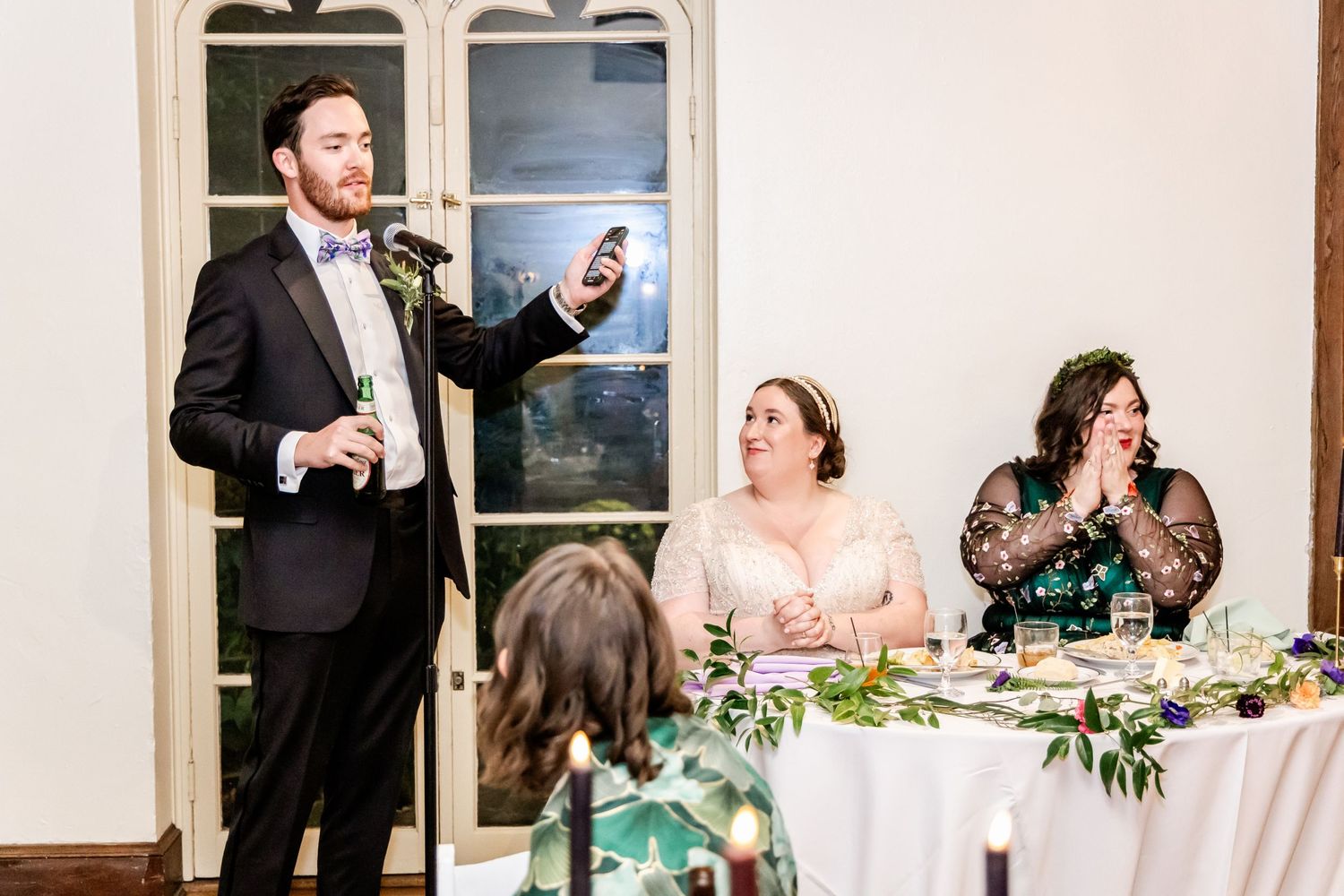 A wedding reception scene with guests seated at decorated tables while someone gives a toast.