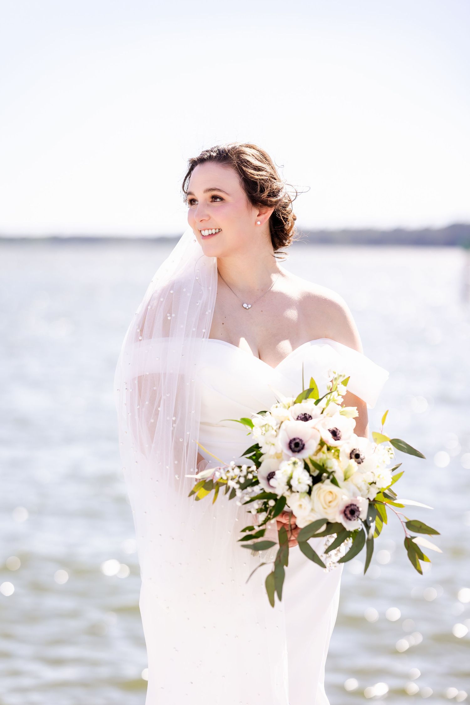 Bride in white wedding dress and delicate shawl smiles while holding elegant white anemone and greenery bouquet near water.