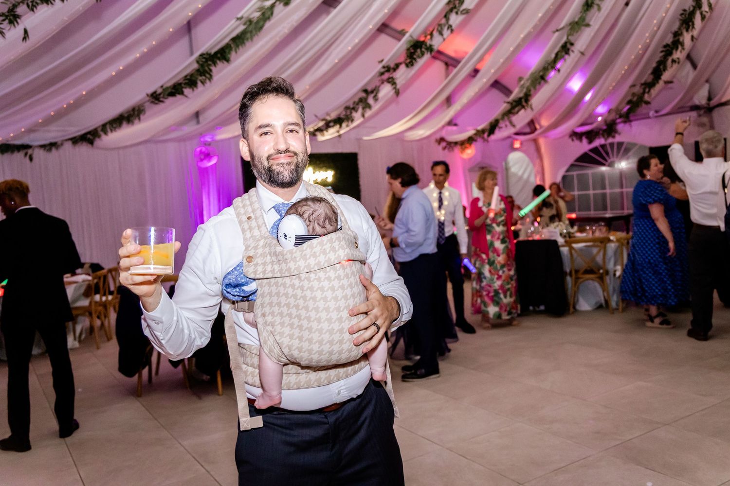 Wedding guests dancing and celebrating at a tent reception with purple ambient lighting.