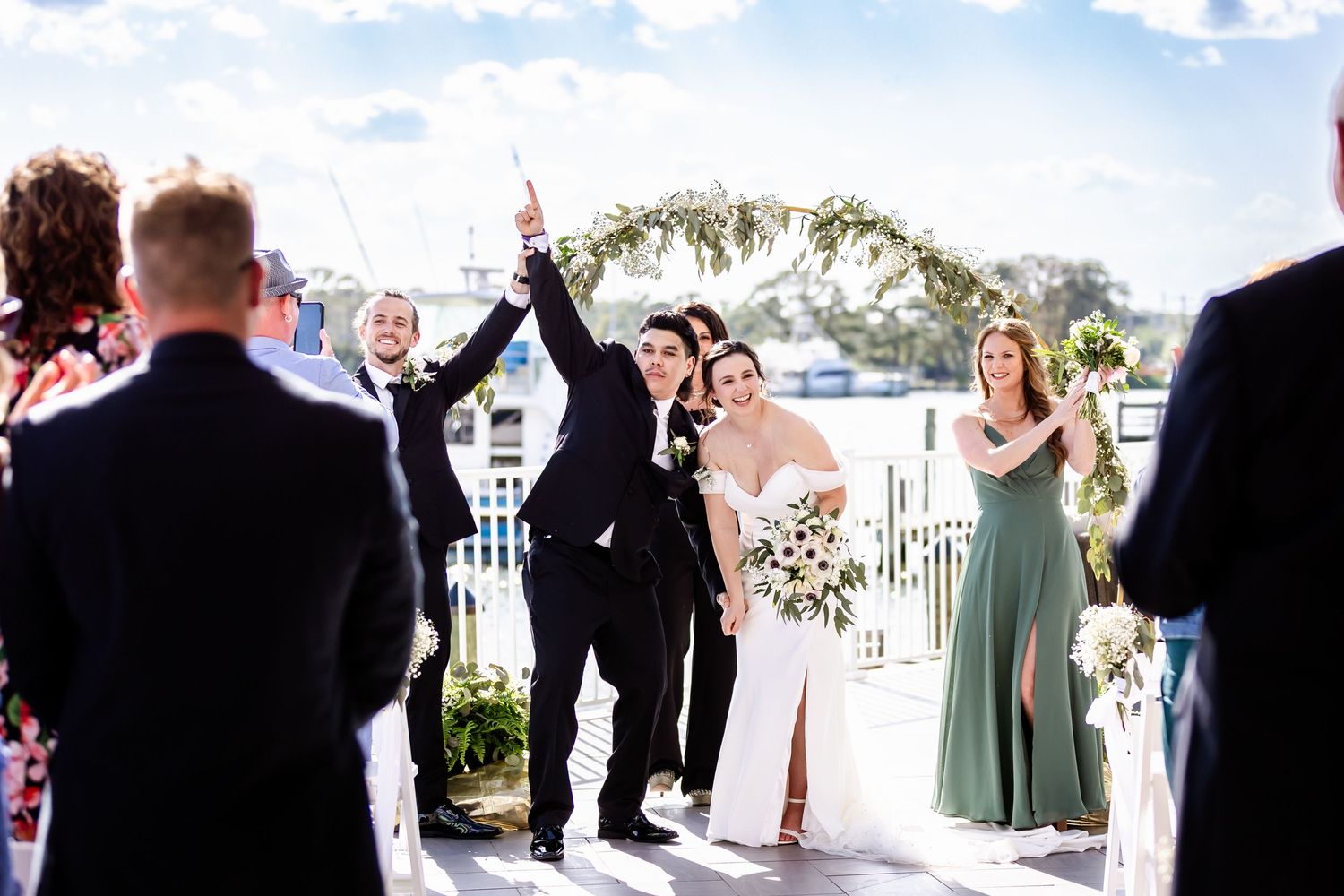 Wedding ceremony celebration with couple and guests cheering under floral arch at outdoor venue during bright sunny day.