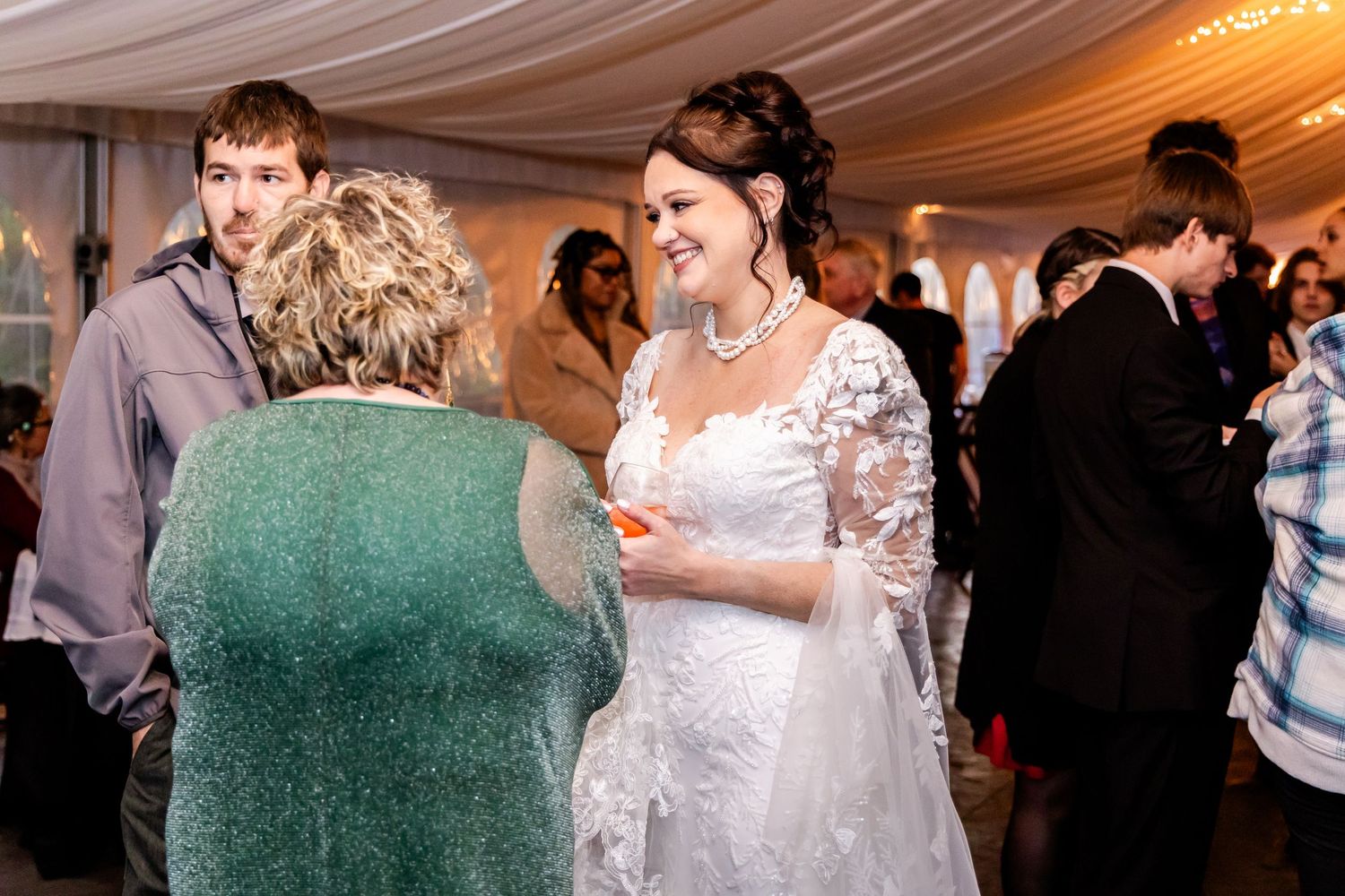 Wedding reception guests mingling under elegant white tent draping and chandeliers.