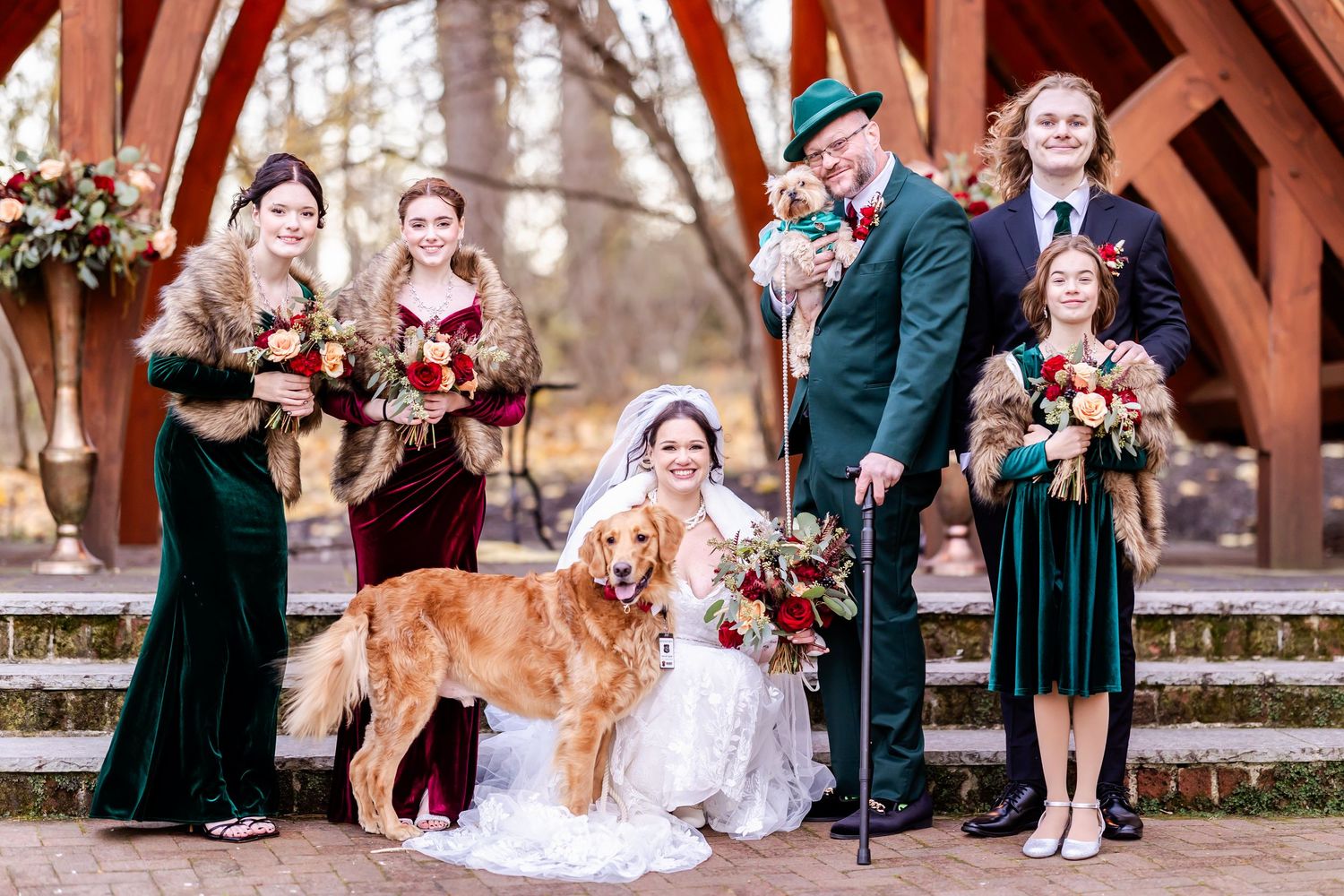 Wedding party poses outdoors with golden retriever dog wearing floral collar at rustic venue.